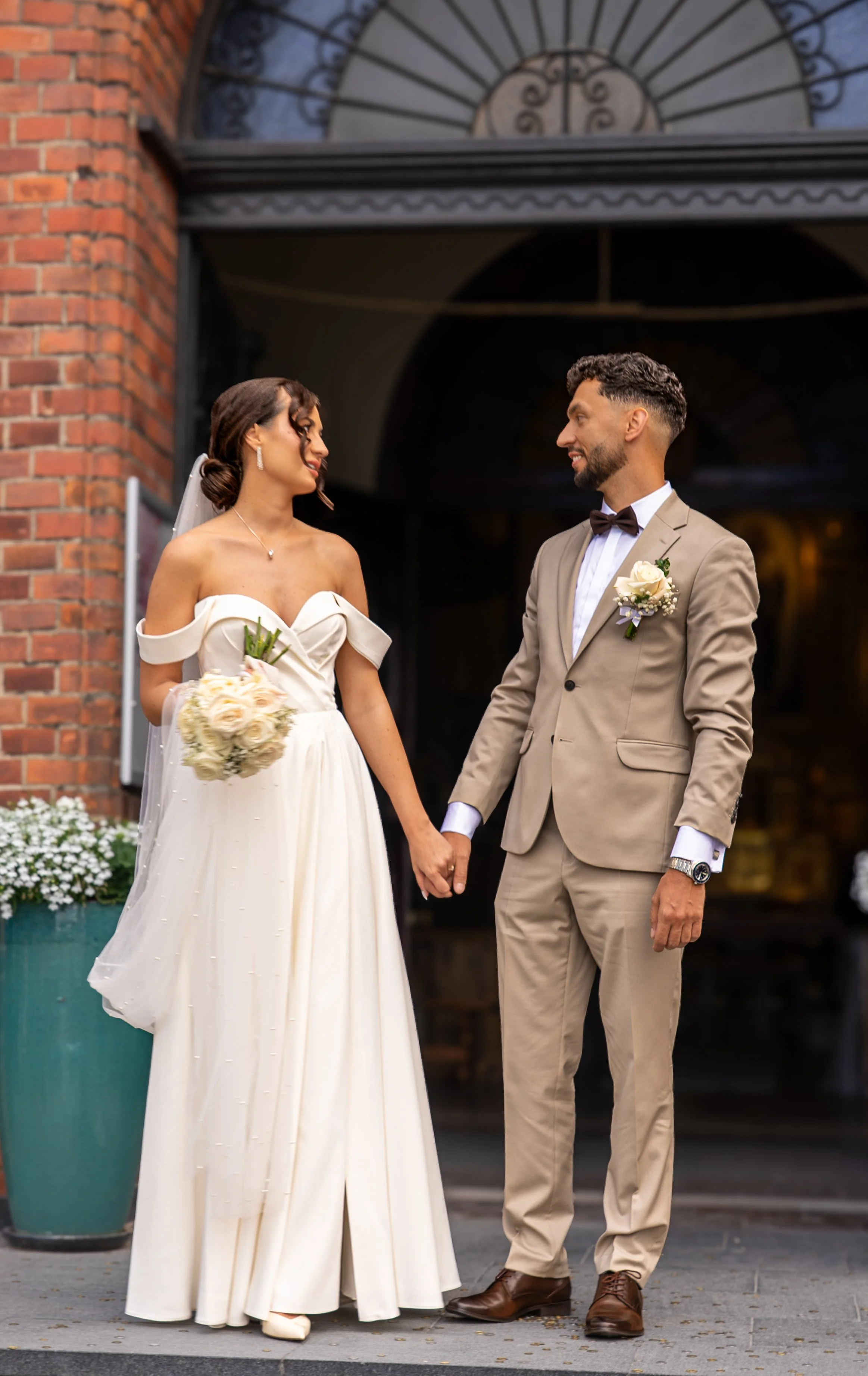 Bride and groom holding hands outside a building at sunset, looking at each other, bride in a white off-shoulder wedding dress holding a bouquet, groom in a beige suit with a bow tie and white shirt.