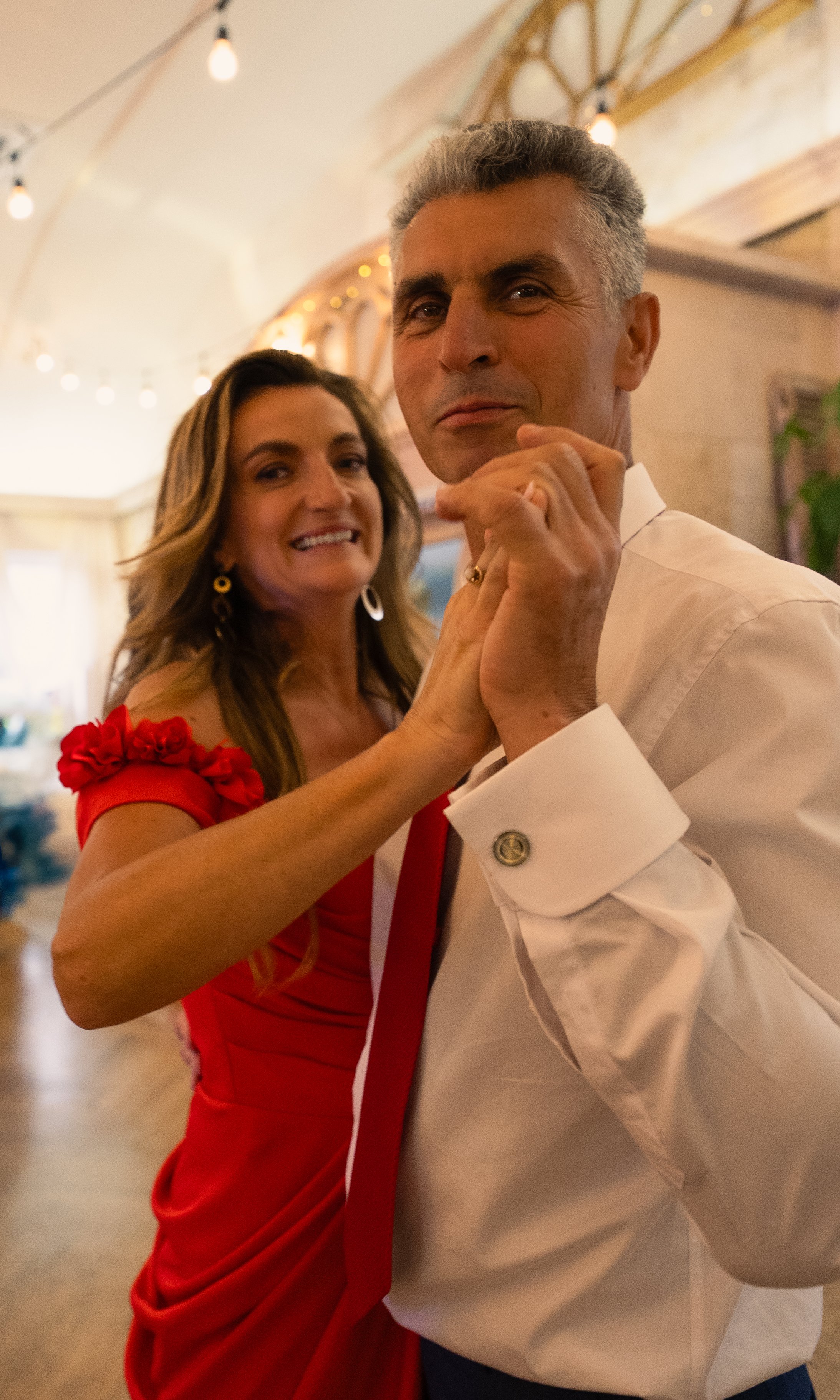 A couple dancing closely together indoors, with the woman wearing a red dress and the man wearing a white shirt.