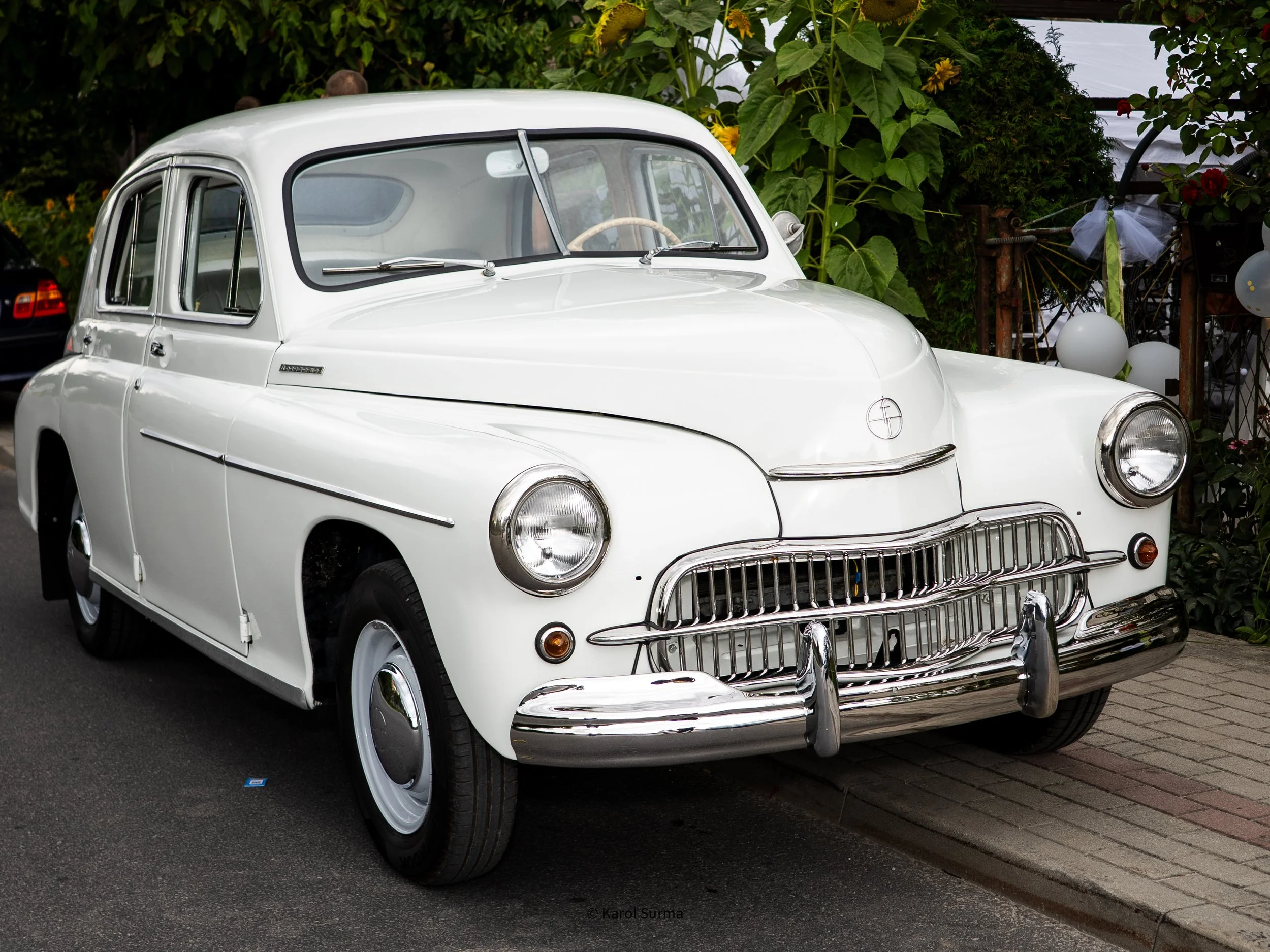 A vintage white car parked on a street with greenery and balloons in the background.