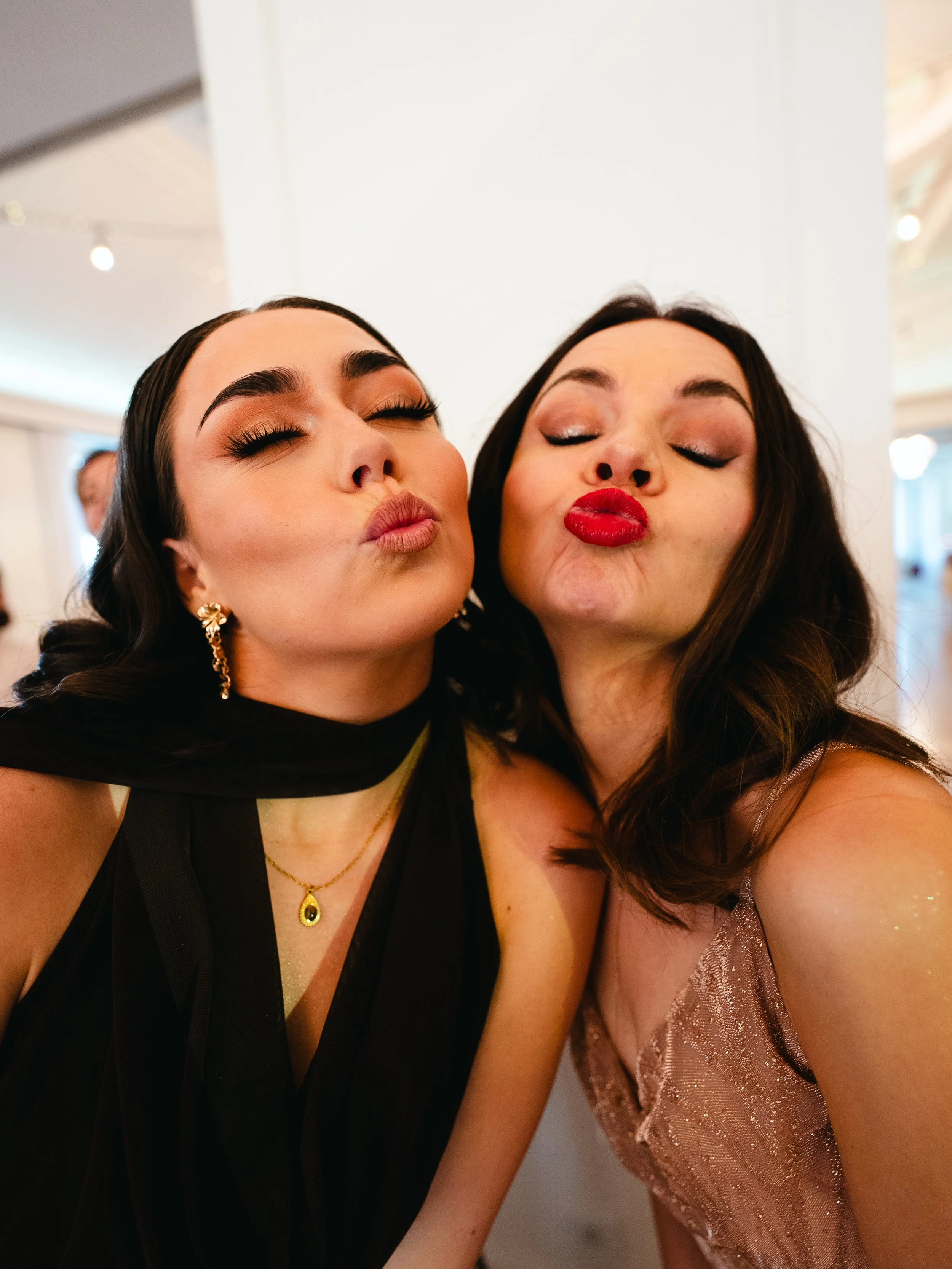 Two women with makeup and jewelry kissing and puckering their lips at an indoor event.