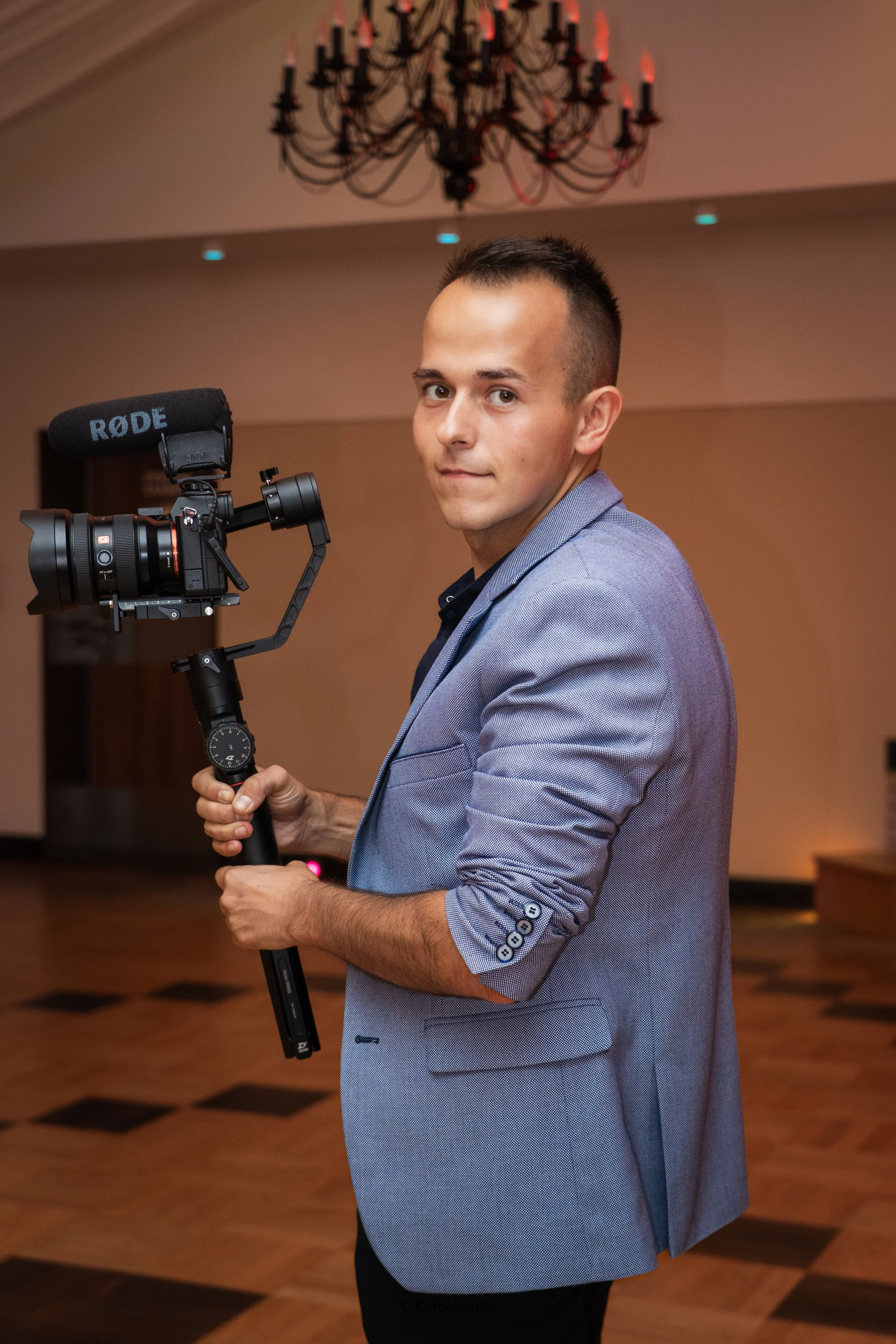 A man in a light blue blazer holding a camera stabilizer with a professional video camera; chandelier hanging from the ceiling in background.