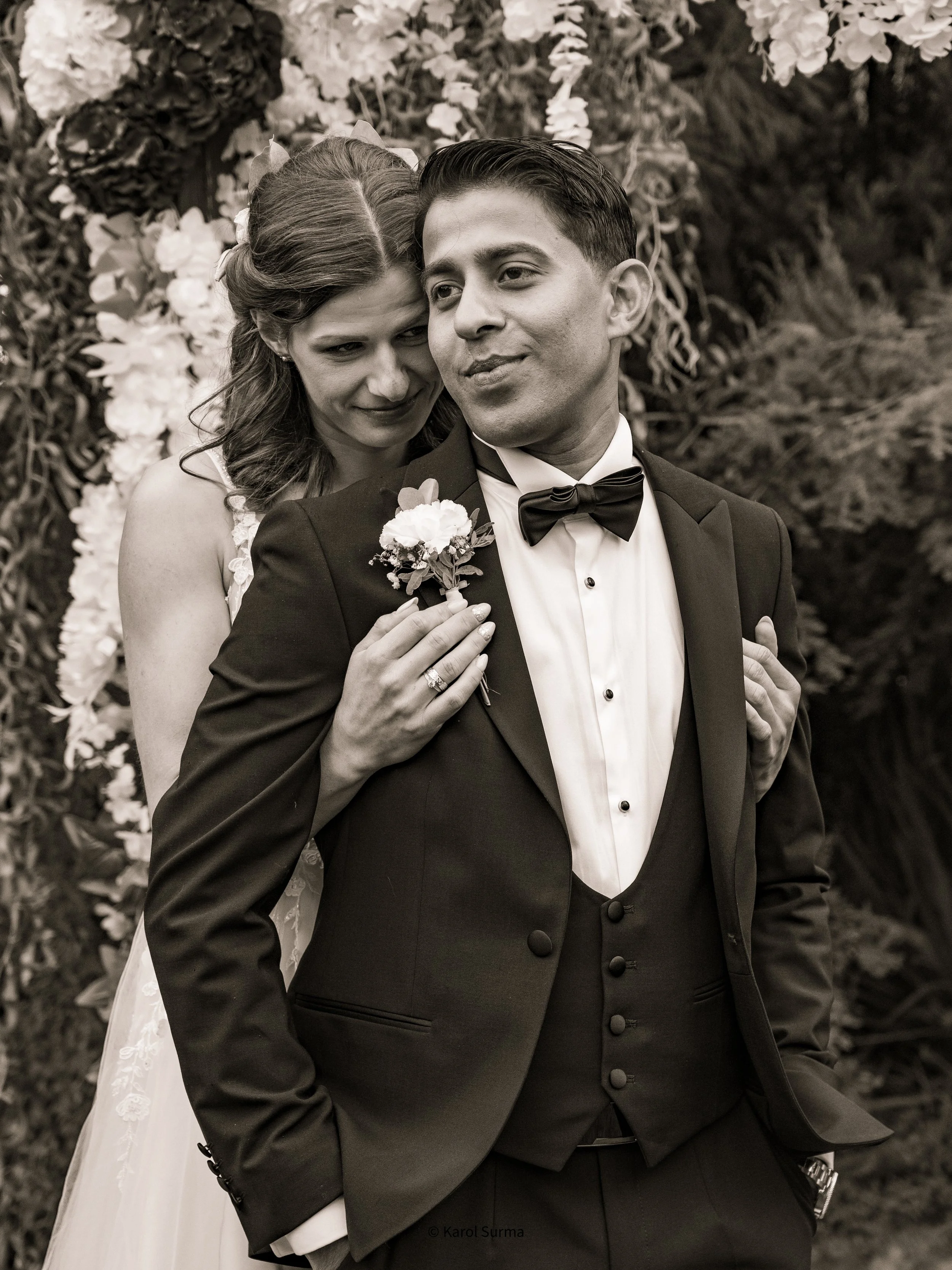 A black and white photo of a wedding couple outdoors. The woman, with wavy hair, stands behind the man, leaning her head on his shoulder and holding his lapel. The man, wearing a tuxedo with a bow tie, stands with his hands in his pockets, and a flower boutonniere on his lapel. They are surrounded by flowers and greenery.