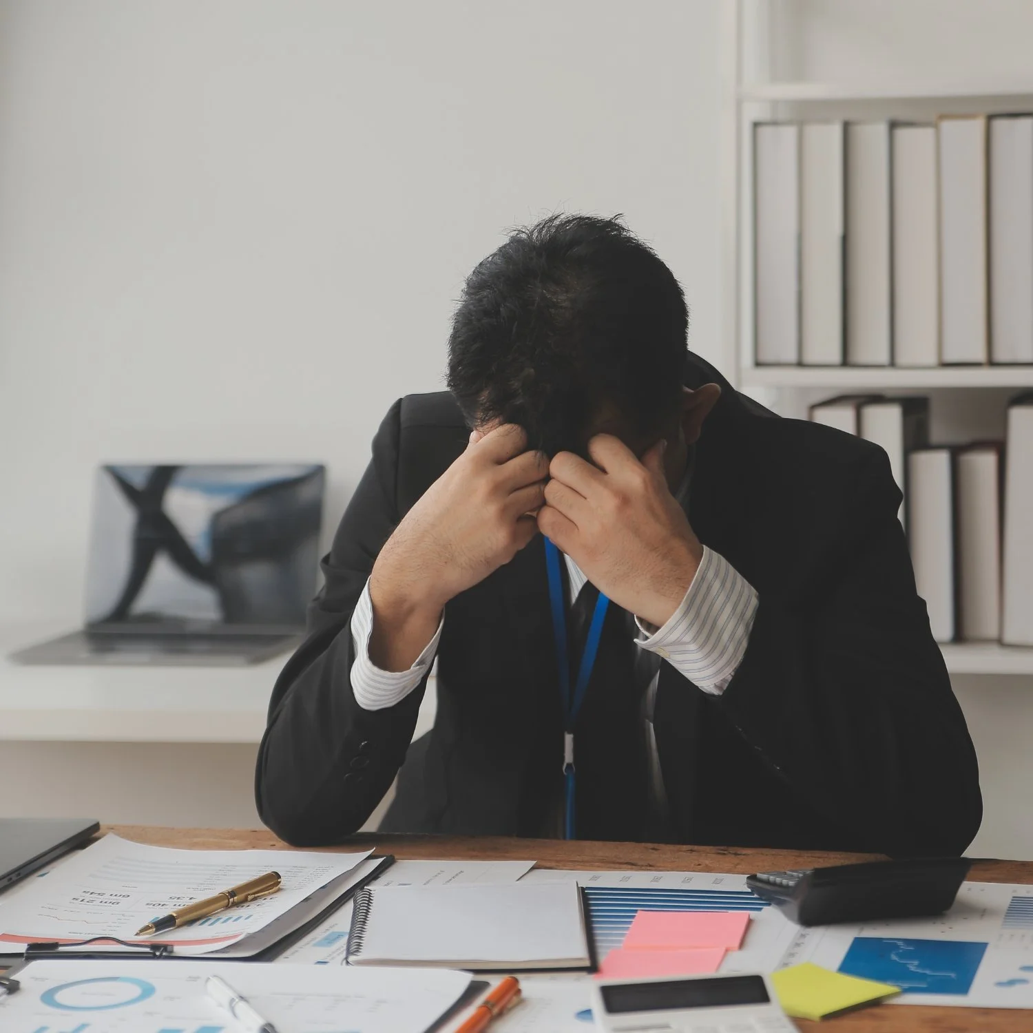 Businessman sitting at a cluttered desk, holding his head in frustration, with papers, a laptop, and office supplies in front of him.