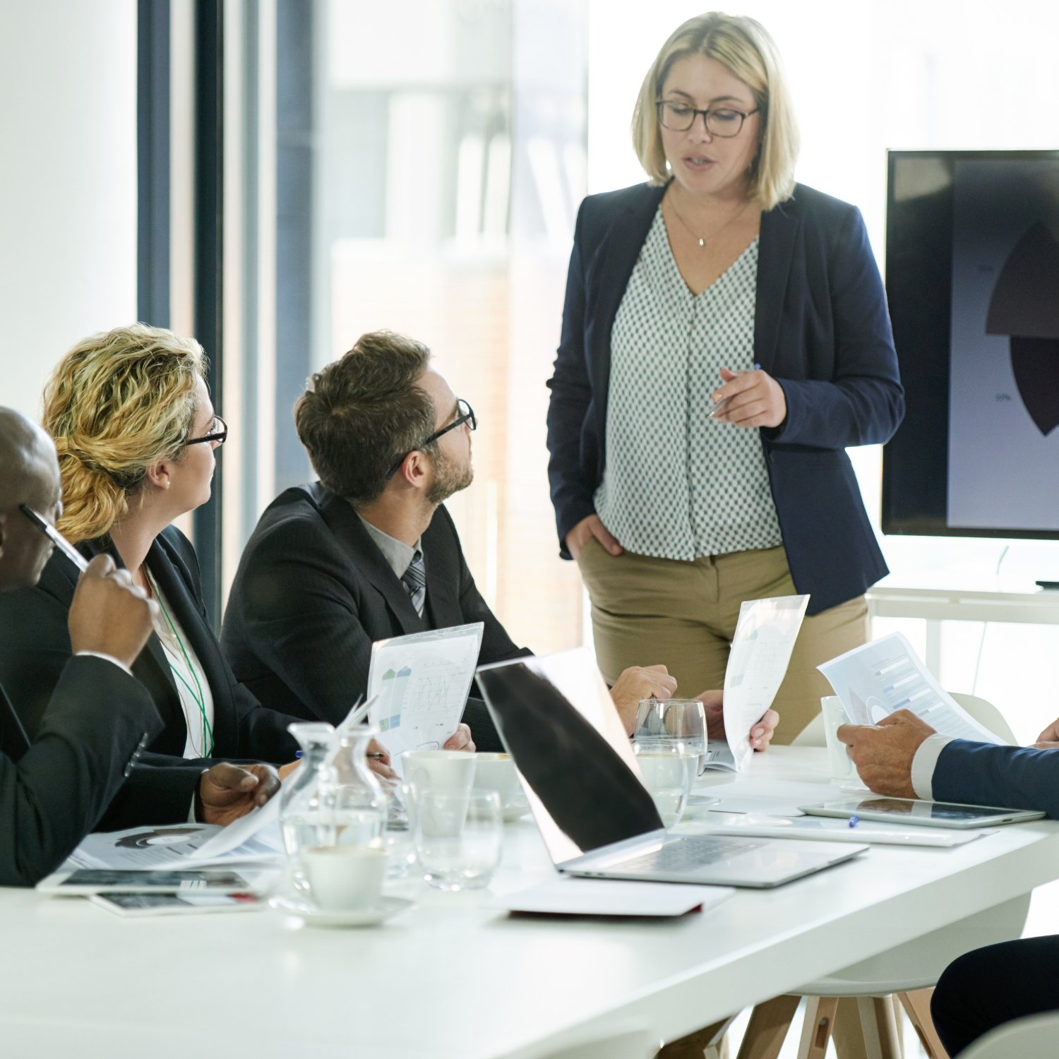 Business meeting with diverse group of women and men in a conference room, woman presenting in front of screen, documents and laptops on table.