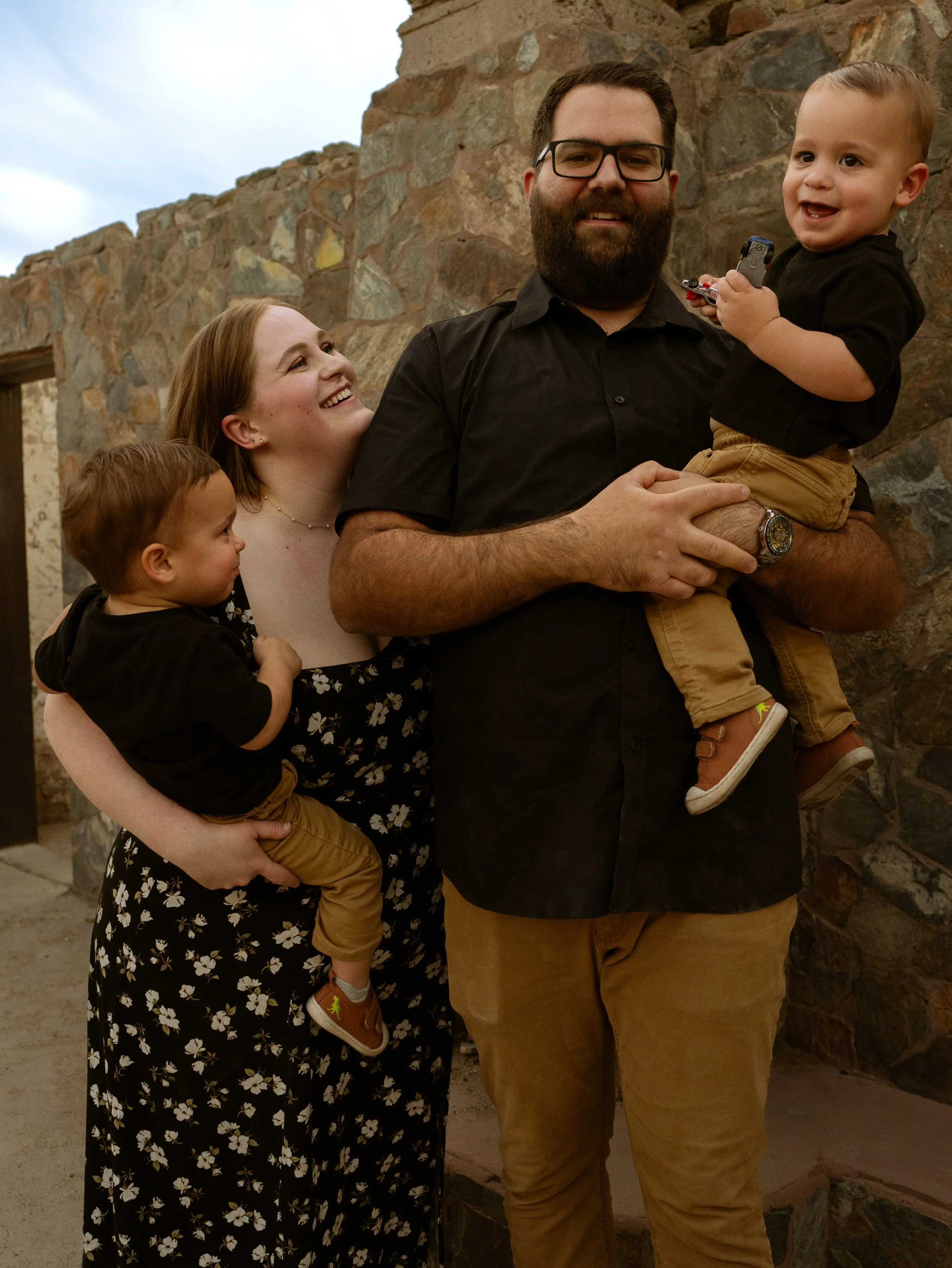 Family of four smiling outdoors near a stone wall, with two young children being held and a woman looking at a man.