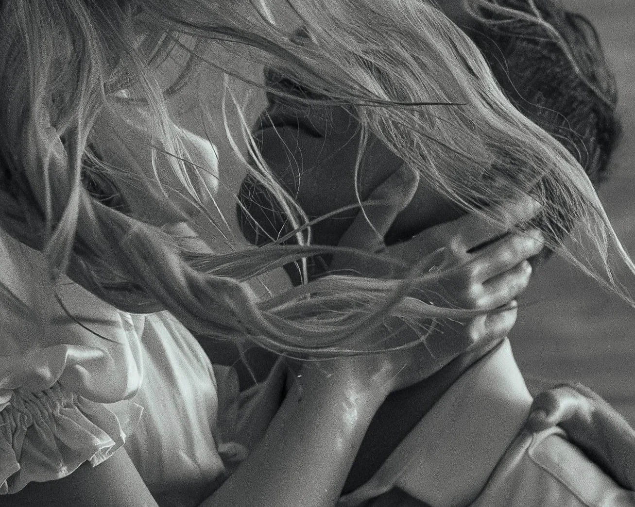 A black and white photo of a young girl with long hair holding her face in her hands, with her eyes closed and head tilted down.