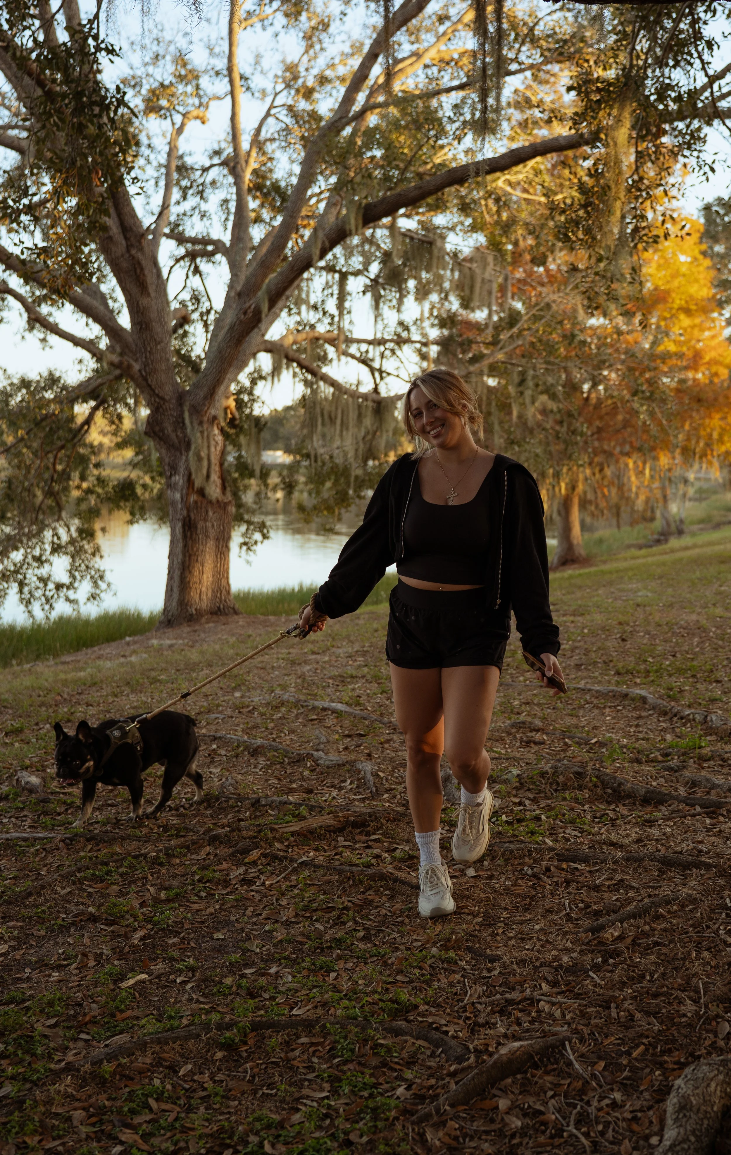 A young woman with blonde hair smiles while walking her small black and tan dog on a leash through a wooded area during sunset. The background features large trees with autumn-colored leaves and a calm body of water.