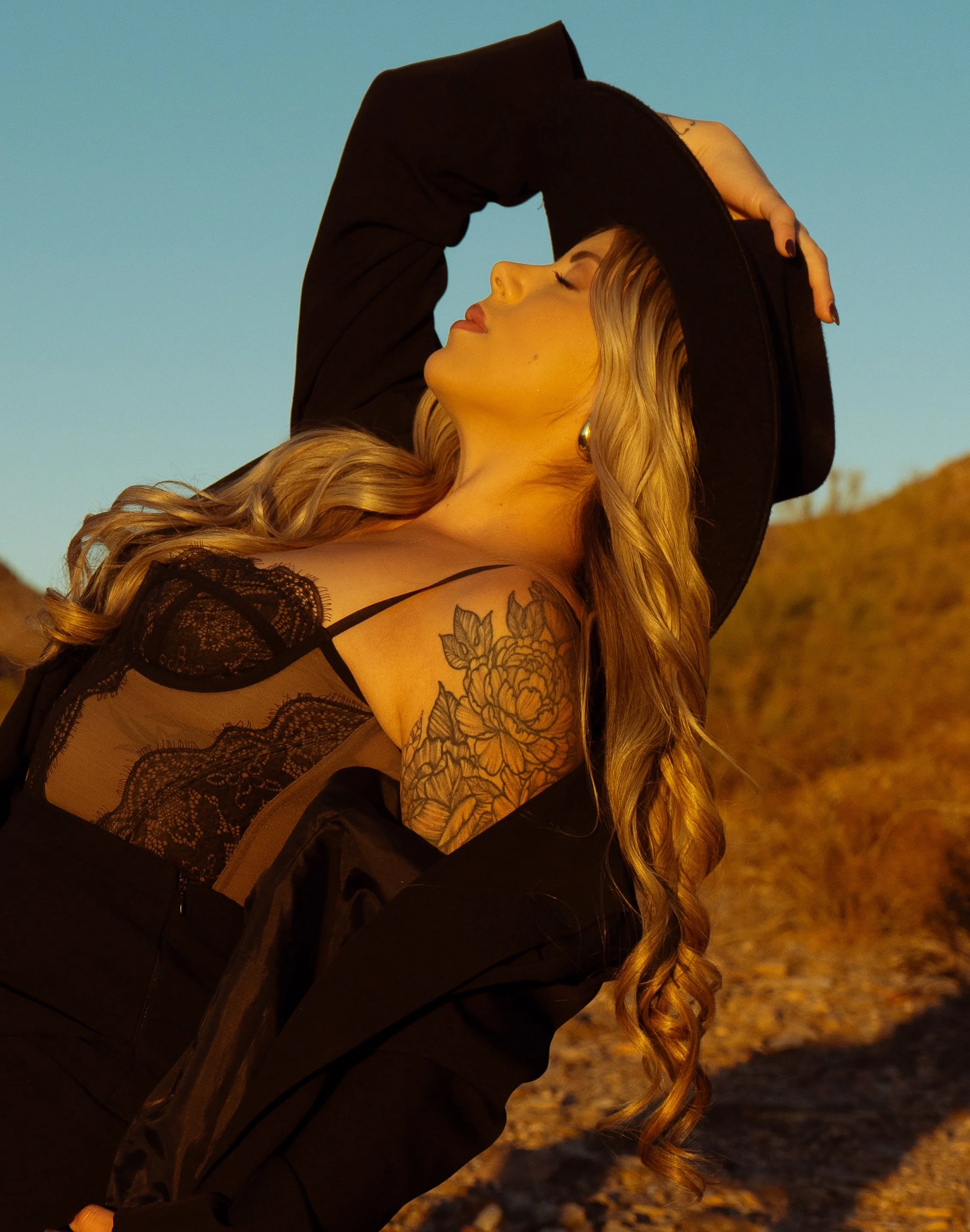 A woman with long wavy hair and a floral tattoo on her shoulder, wearing a black lace top and a black jacket, holding a wide-brimmed hat on her head, standing outdoors during sunset.