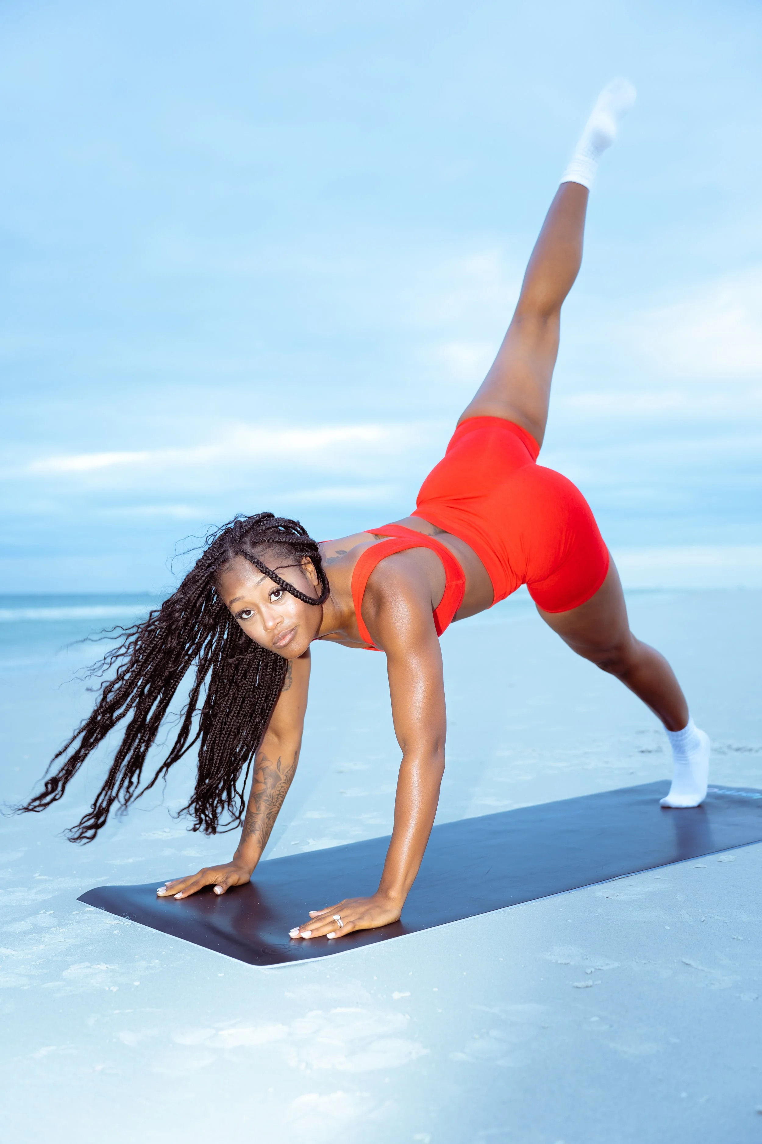 A woman in red athletic wear practicing yoga outdoors on a blue yoga mat with a cloudy sky in the background, performing a side plank pose.