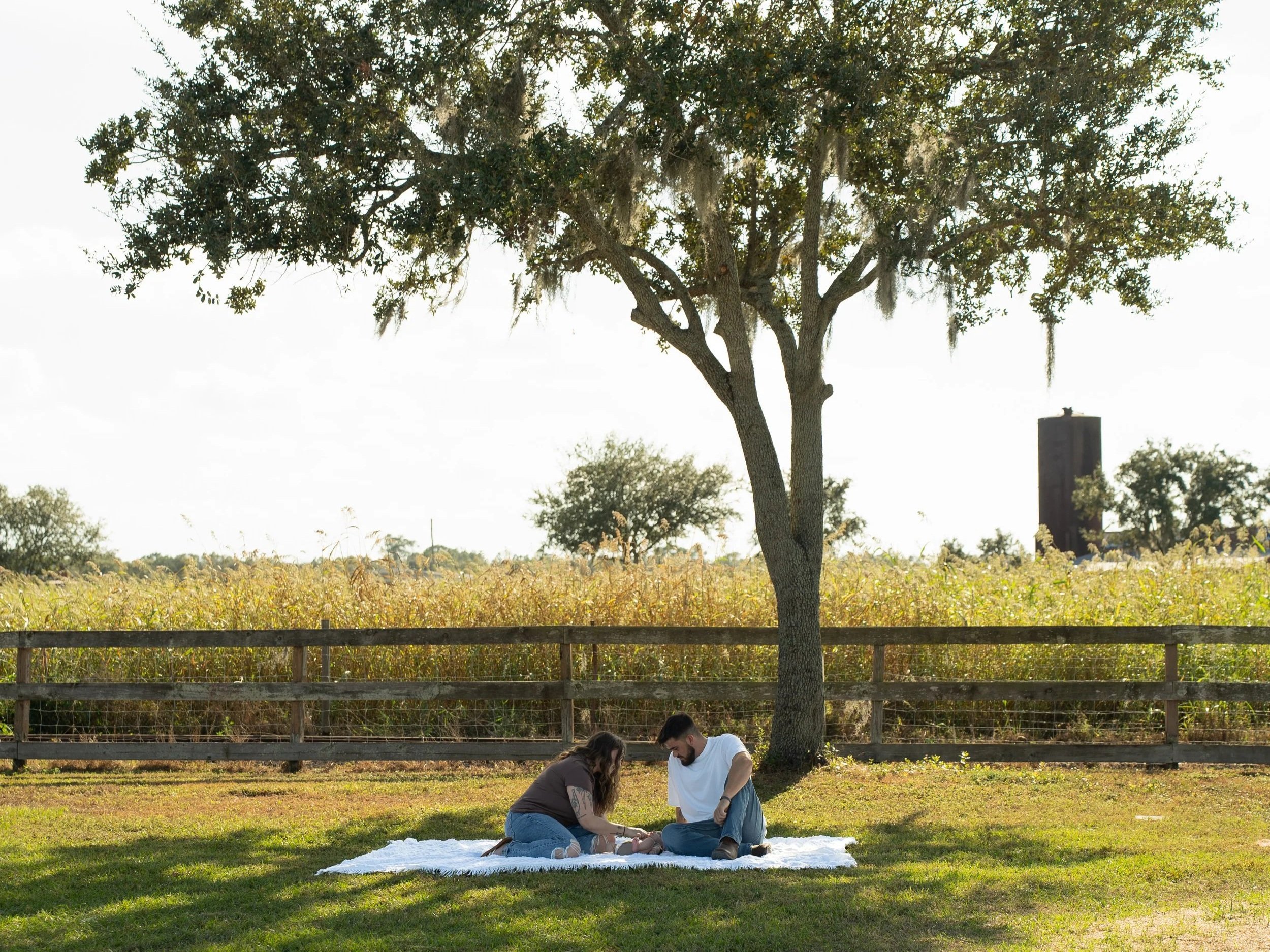 A couple kneeling on a white blanket on grass, participating in a prayer or blessing ceremony outdoors, with trees and a fence in the background.