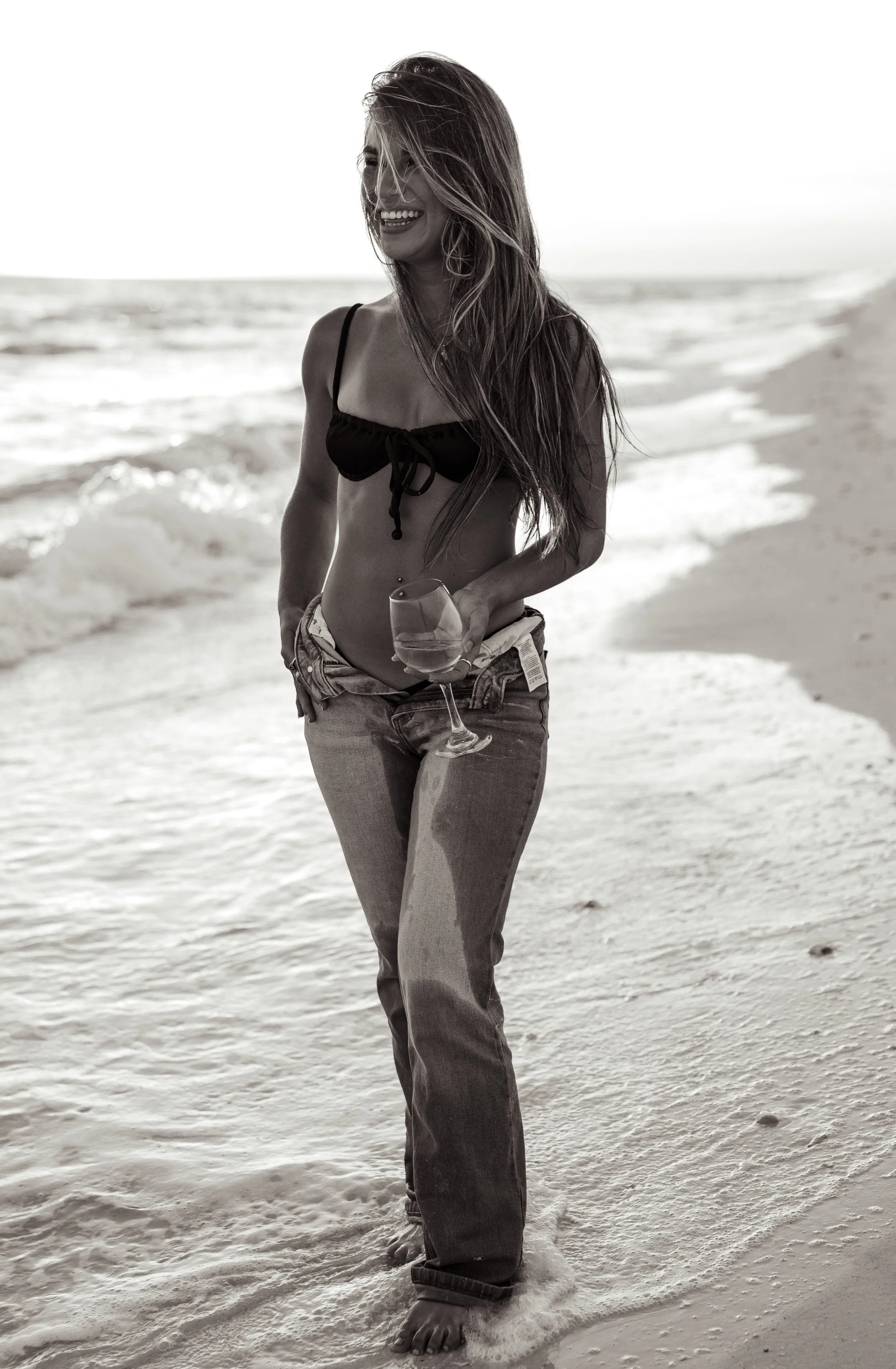 A woman with long hair smiling and holding a wine glass at the beach, wearing a black bikini top and jeans, with waves in the background.