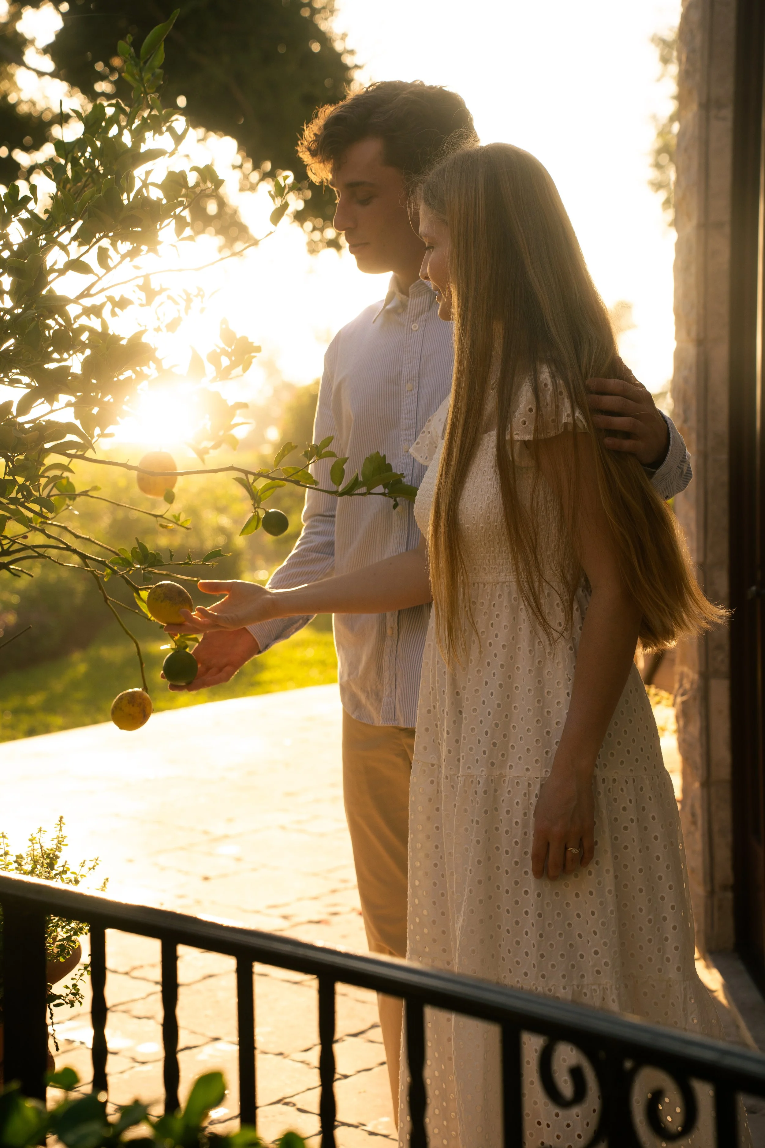 A couple standing outside near a fence during sunset, with the woman holding a fruit from a tree.