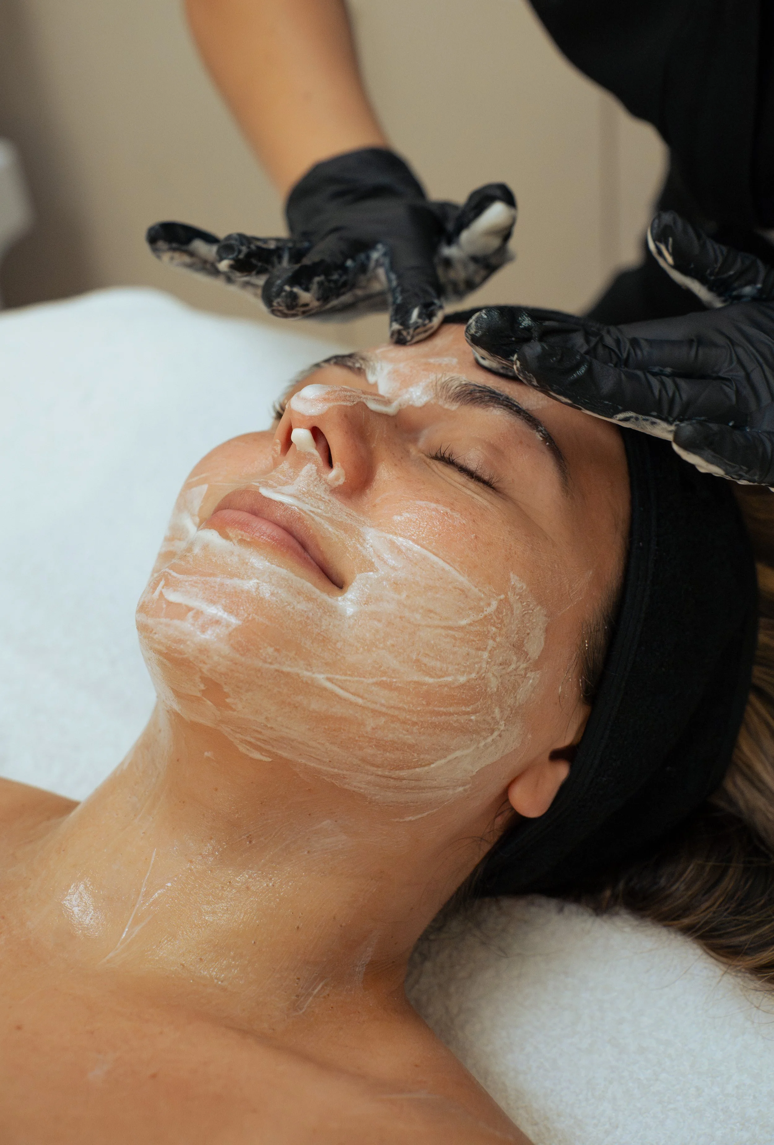 A person with closed eyes lying on a surface is receiving a facial treatment, with foam applied to their face, while a technician wearing black gloves and black clothing applies the foam.