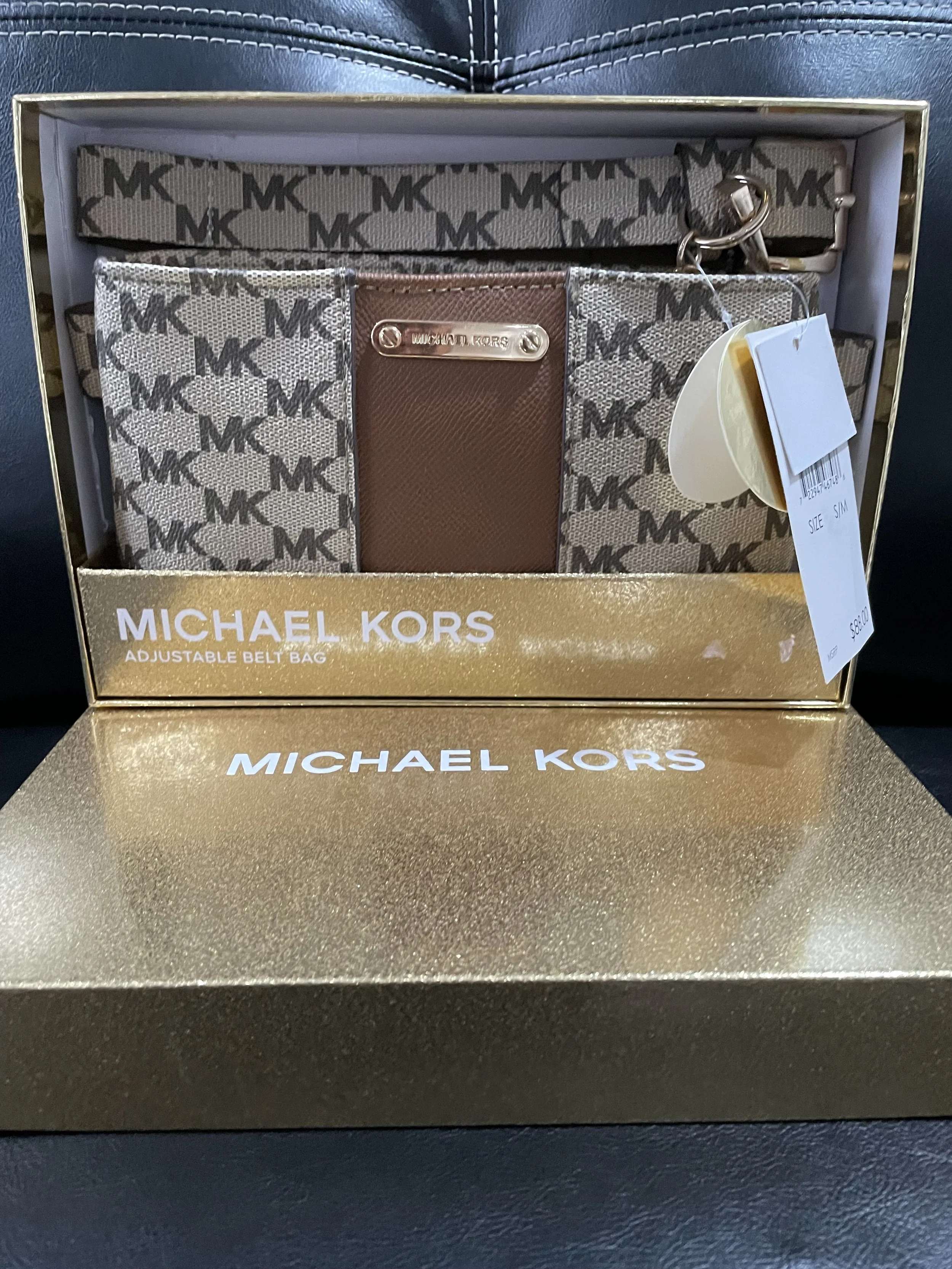Set de cinturón, cartera y llavero de la marca Michael Kors en caja dorada con el logo. La cartera tiene un patrón con letras MK y una etiqueta metálica.