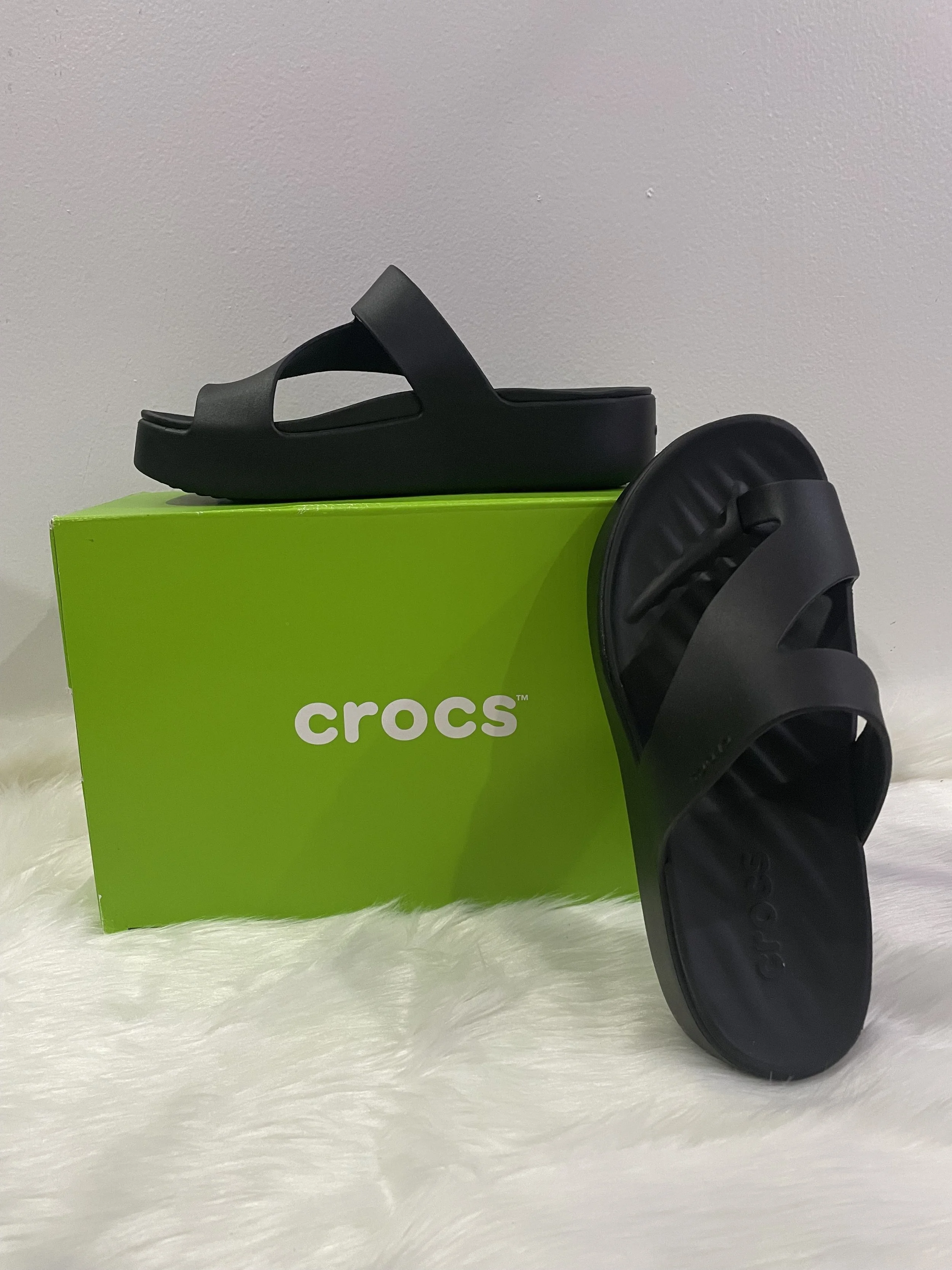 Sandalias negras de la marca Crocs sobre una caja verde de Crocs y una superficie de peluche blanco.