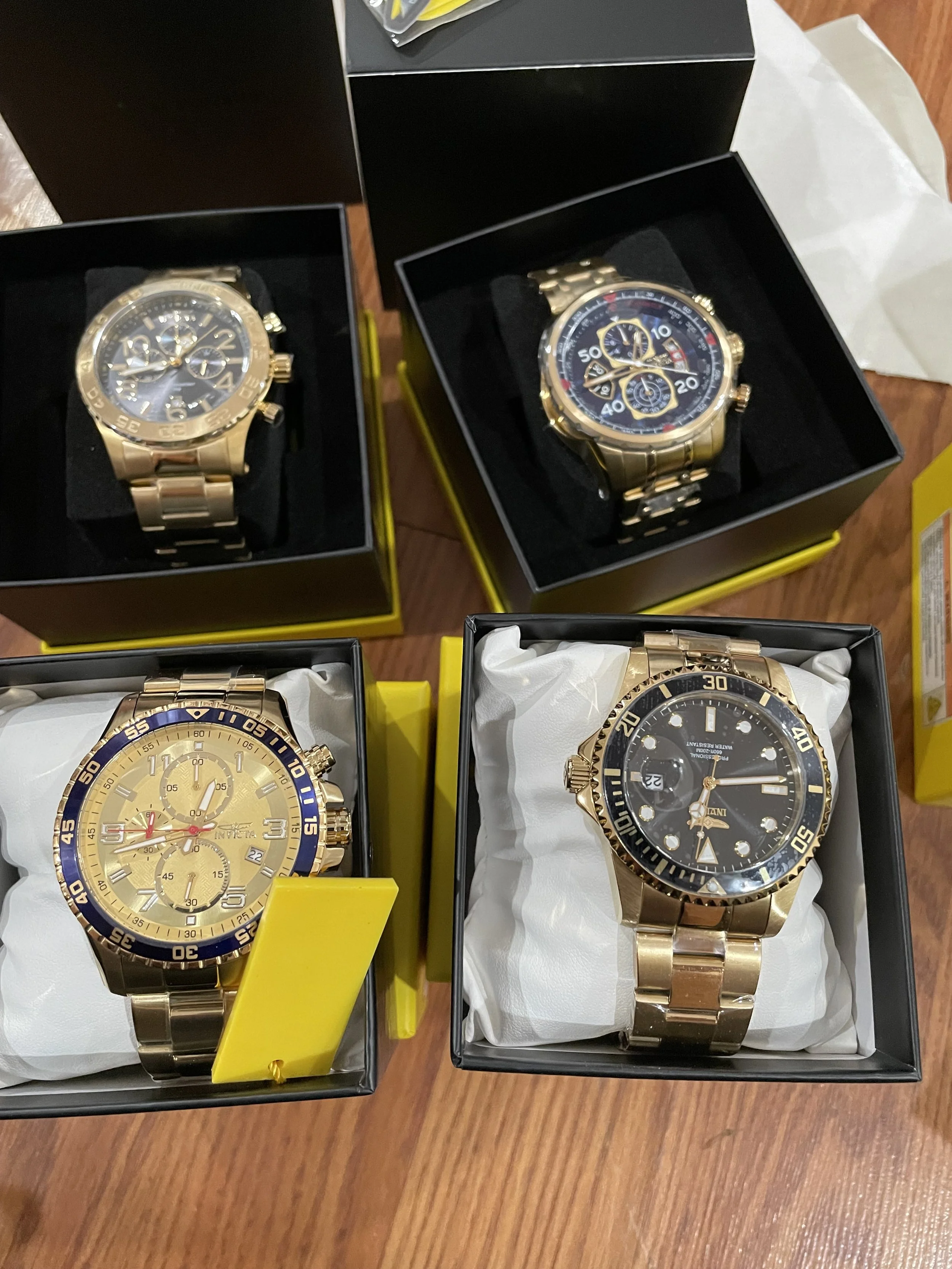 Cuatro relojes de pulsera en cajas, dos con cajas negras y amarillas y dos con cajas negras y blancas, todos con correas de metal dorado, exhibidos sobre una mesa de madera.