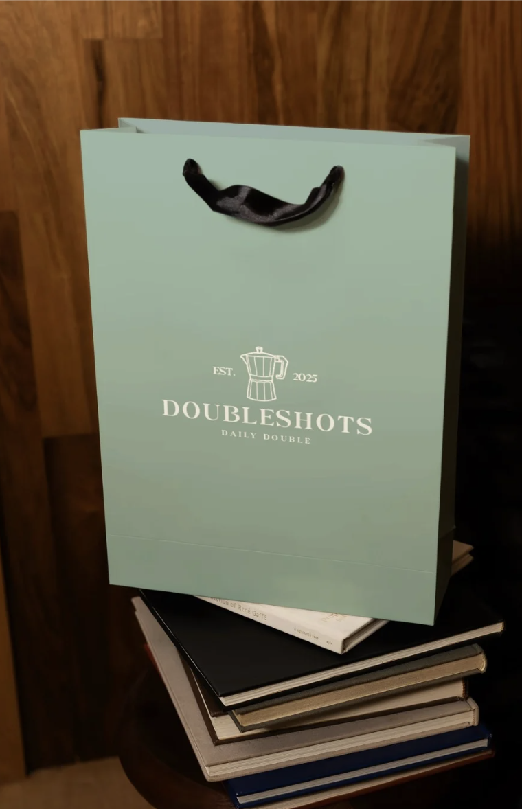 A light green shopping bag with black handles sitting on top of a stack of books. The bag has a logo with a moka pot and text that reads "Est. 2025 DoubleShots Daily Double."