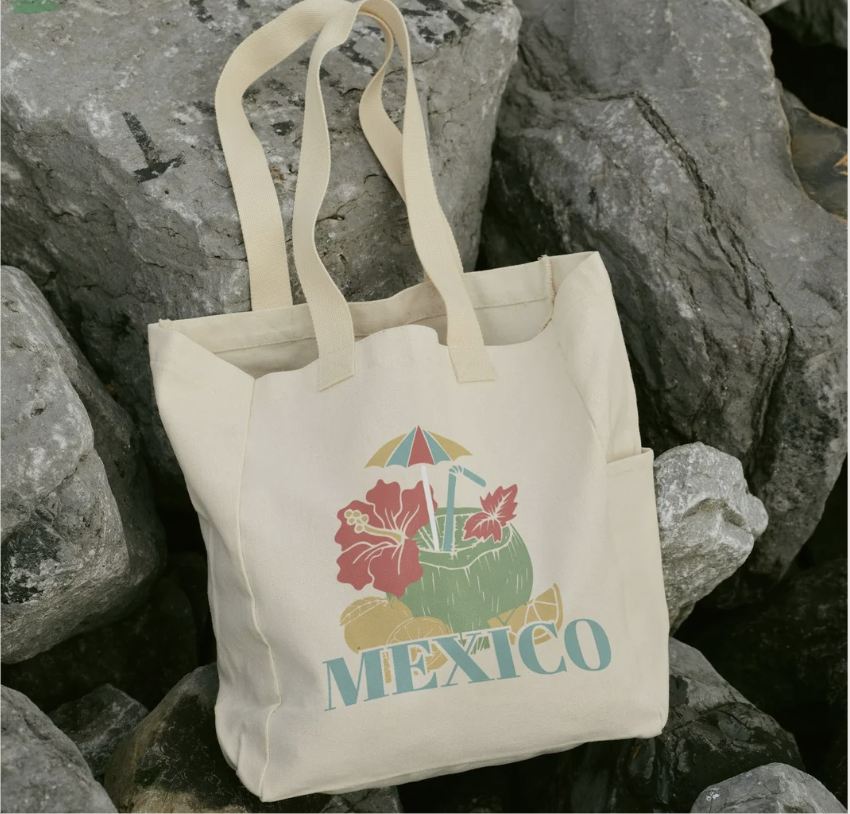 A beige tote bag with the word "Mexico" in large blue letters. The design features a cocktail with a straw, umbrella, hibiscus flowers, and tropical elements, set against a background of rocks.