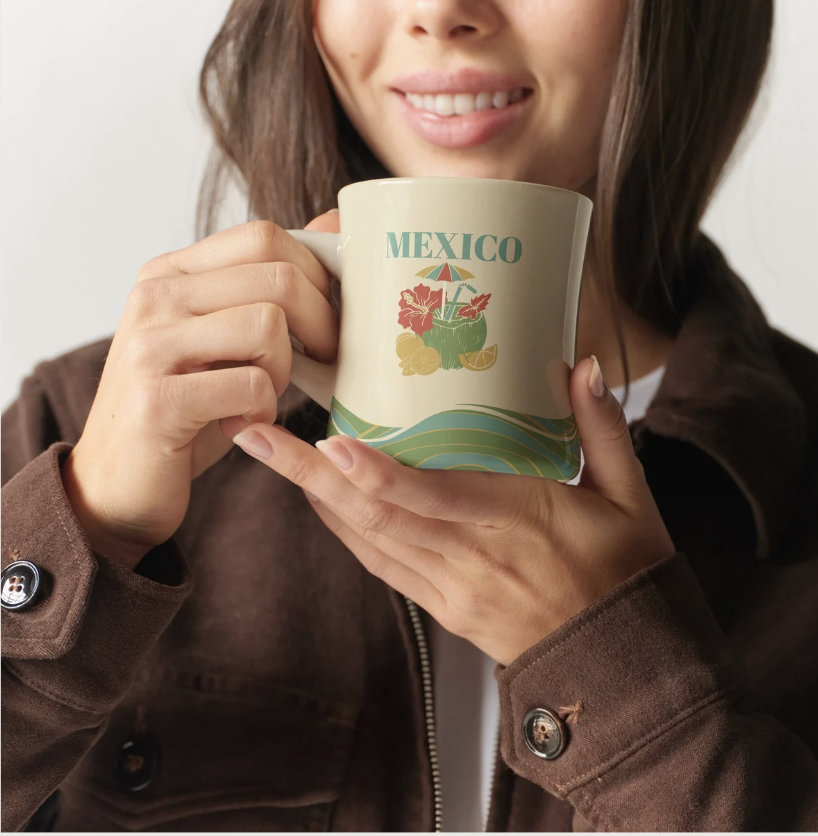 A woman holding a mug with a Mexico design, featuring hibiscus flowers, an umbrella, lemons, and ocean waves, and smiling.