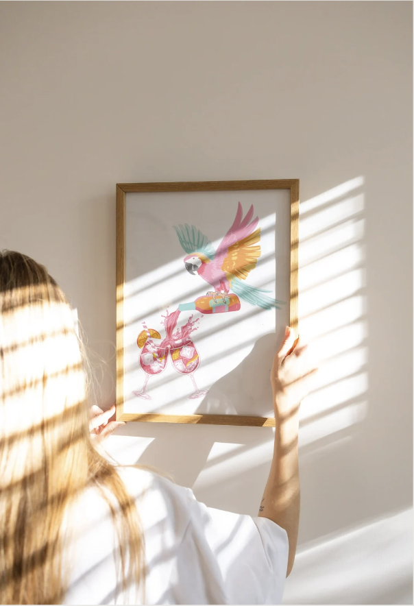 Person holding a framed art print of a colorful parrot standing on a purse with the background of a plain off-white wall with sunlight and window blinds casting shadows.
