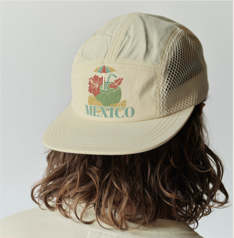 A person with curly brown hair wearing a beige cap with an embroidered design of a coconut drink with a straw, an umbrella, hibiscus flowers, and the word "Mexico" on it.