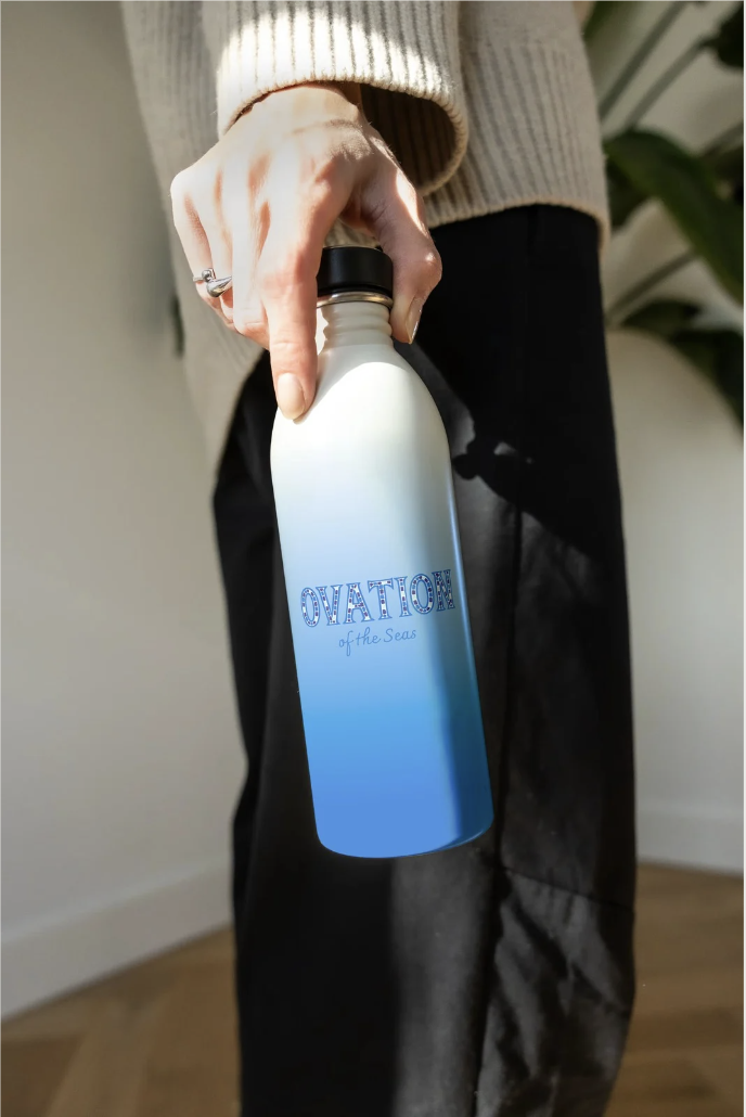 A person holding a white water bottle with a black lid, featuring a blue gradient design and text that reads 'OVATION of the Seas.' The person is wearing a beige sweater and black pants, standing indoors with a plant in the background.