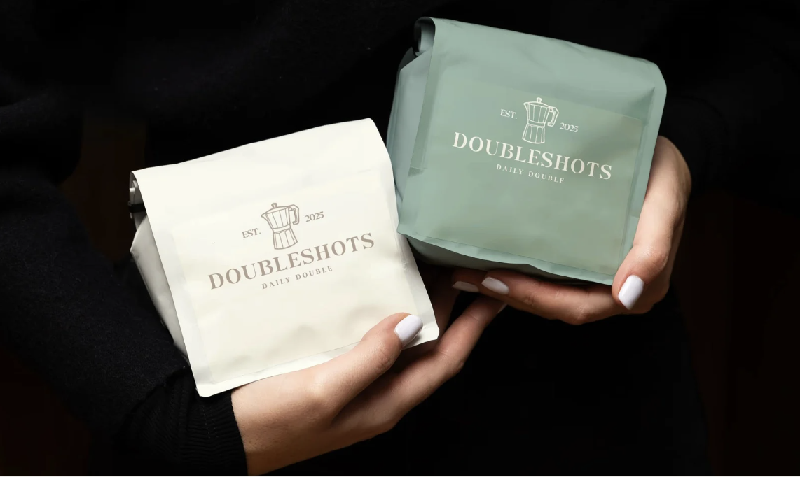 Two hands holding packages labeled "DoubleShots Daily Double," with one package white and the other light green, featuring a coffee pot icon and the text "EST 2025."