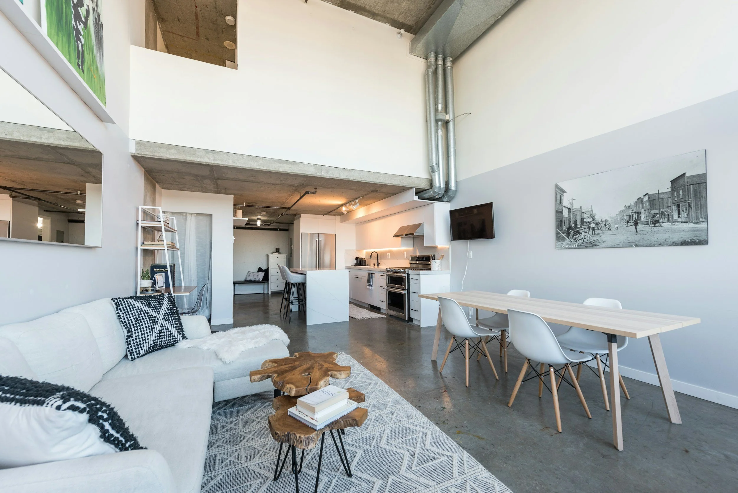 Open living space with white sofa, wooden coffee table, and area rug. Kitchen with white cabinets, stainless steel appliances, and a breakfast bar with black chairs. Dining table with white chairs. Wall art and a mounted flat-screen TV. Exposed concrete floor and ceiling with visible ductwork.