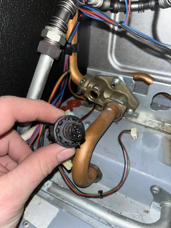 Close-up of a person's hand holding a circular electrical component inside an appliance, surrounded by wires, metallic pipes, and internal parts.