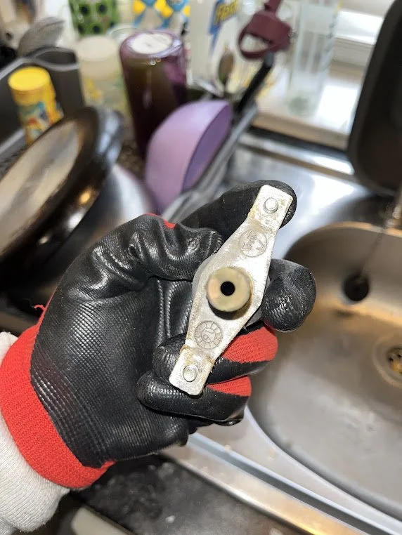 A person wearing a black and red glove holding a small metal part over a stainless steel sink. The background includes various kitchen items like a purple rice cooker, yellow and blue containers, and a window.