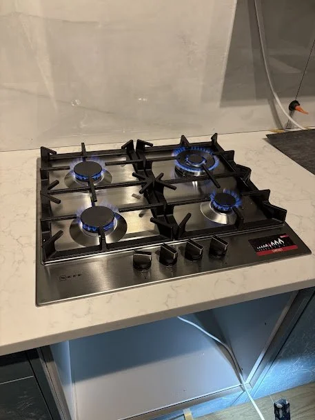 Stainless steel four-burner gas stove with blue flames on three burners, on a white marble countertop.