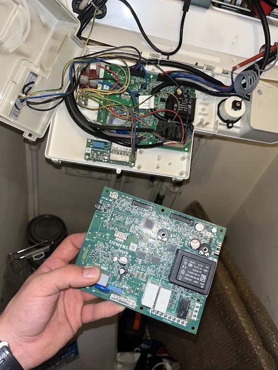 A person holding a circuit board labeled Siemens, showing electronic components, with an open electrical box containing wiring, circuit boards, and other electronic components in the background.