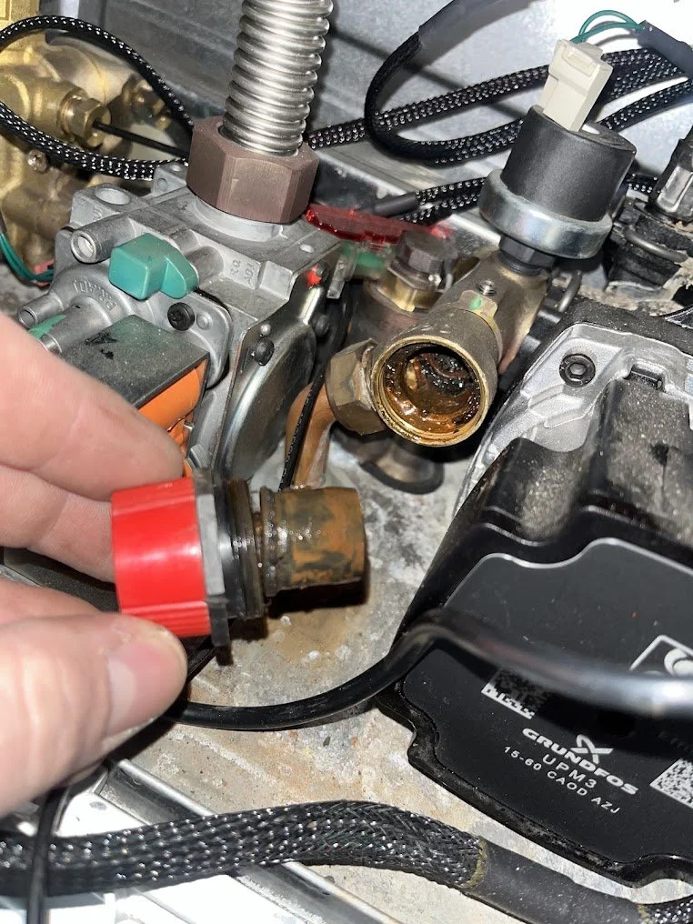 Close-up of a mechanic's hand holding a rusty sensor part near a car engine. The engine has oil and dirt, with exposed pipes, wires, and a focus on a component that appears to be a fuel or oil system part.