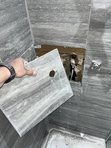 Person holding a gray tile with a hole, installing it on a bathroom wall with gray tiles, exposing plumbing behind the wall.
