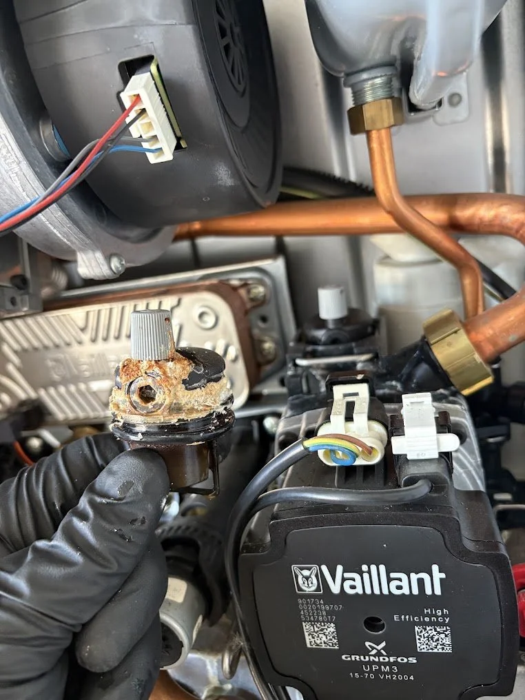 Close-up of a technician's hand holding a corroded and damaged sensor or valve part inside an appliance, with visible copper tubing and electrical connectors nearby.