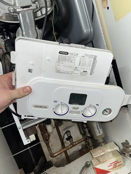 A person holding a white Potterness control panel in front of a plumbing system with pipes and a black component, possibly part of a water heater or boiler, in a utility room.