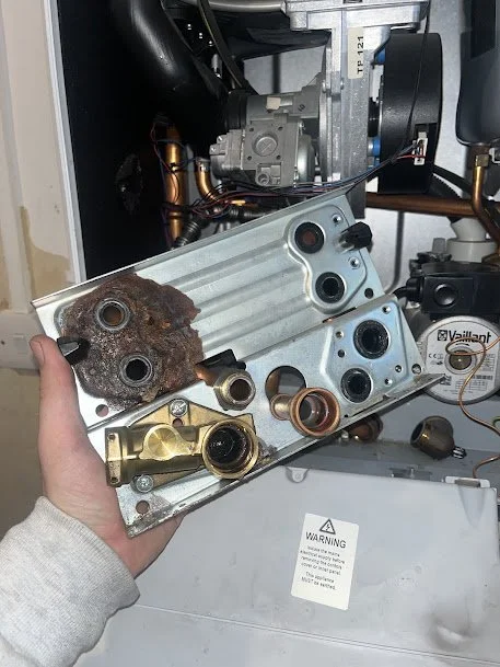 Person holding a burned metallic engine part with rust and oil residue, inside a mechanical or appliance setting with various tools and components nearby.