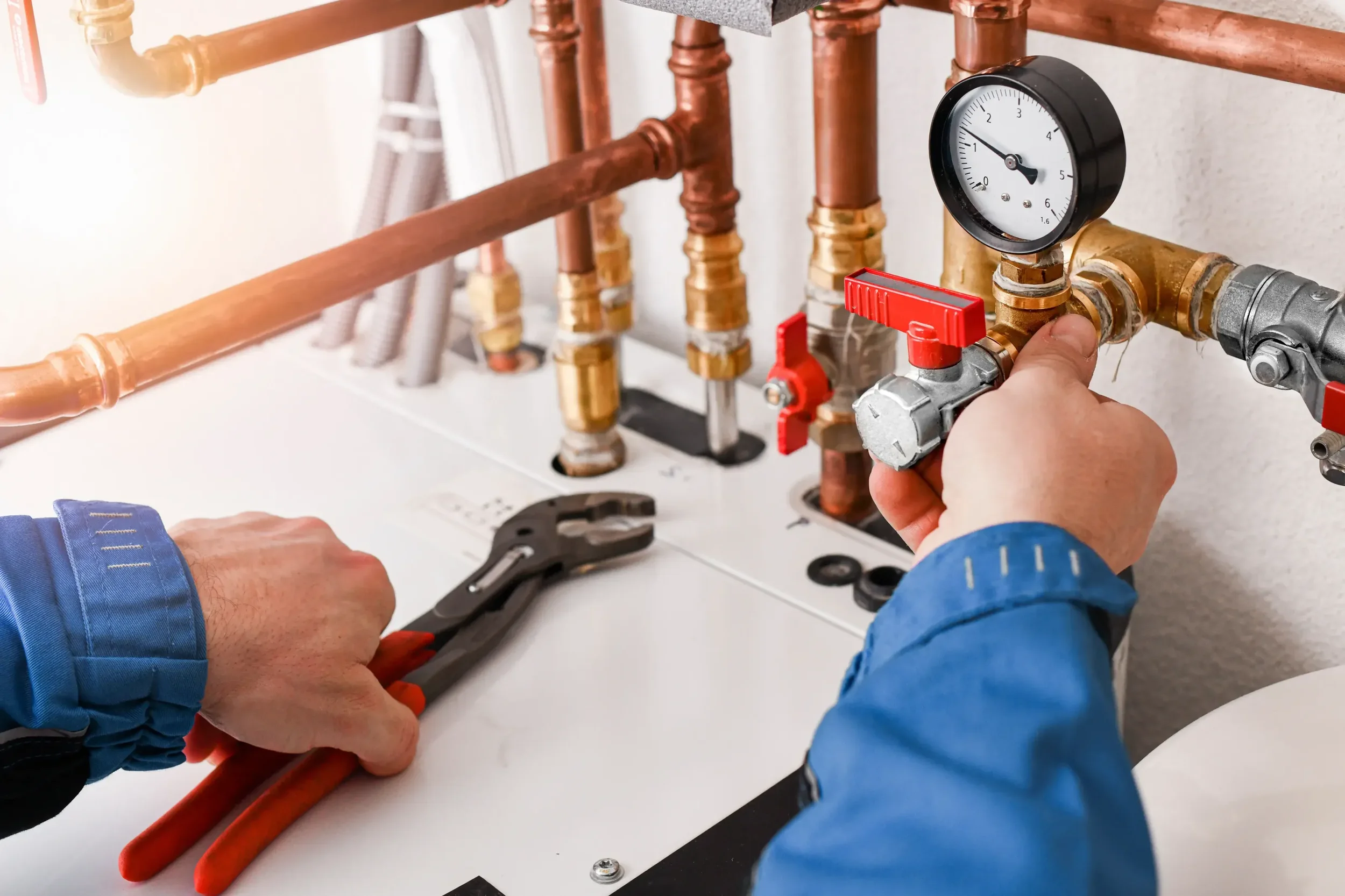 A person wearing a blue jacket is working on copper pipes, using a wrench and adjusting a meter gauge, in a plumbing setup with multiple pipes and valves.