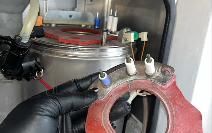A person wearing black gloves holding a circular metal plate with three spark plug wires attached, inside a technical or industrial machine.