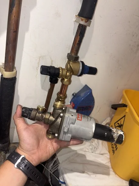 A person's hand holding a plumbing pump with pipes and valves, in a utility room with cleaning supplies and a yellow caution bucket.