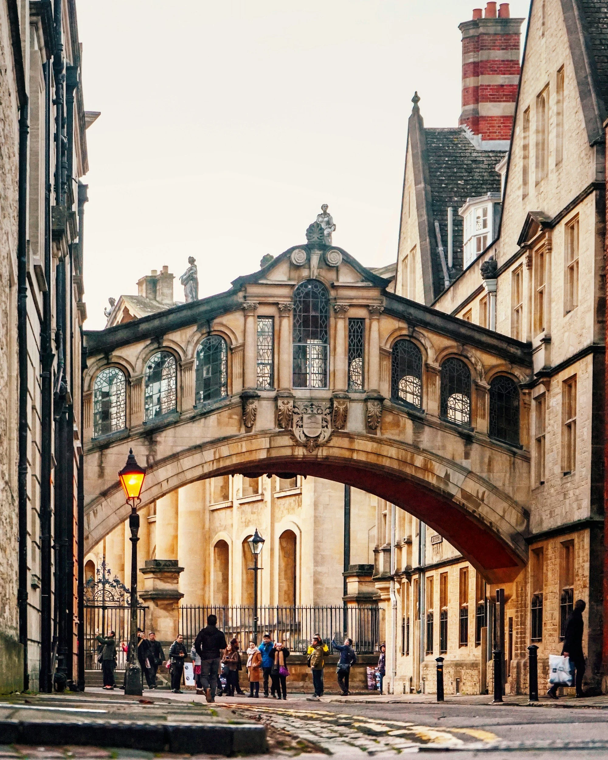 Historical bridge with arched windows connecting buildings in a city street, with pedestrians walking underneath and streetlamps, captured during daytime.