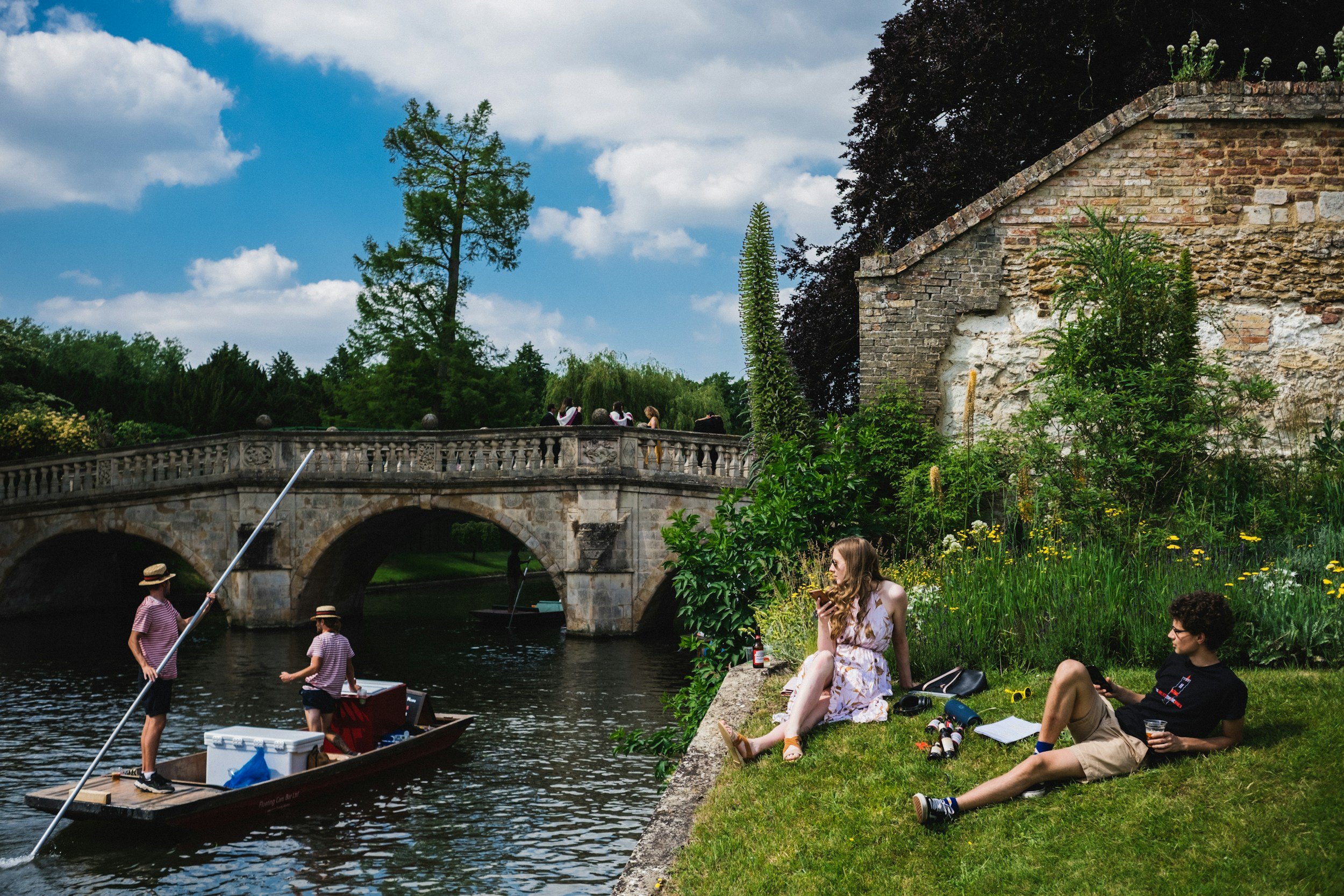 People relaxing on grass near a river with a stone bridge, boat with rowers, and lush greenery.