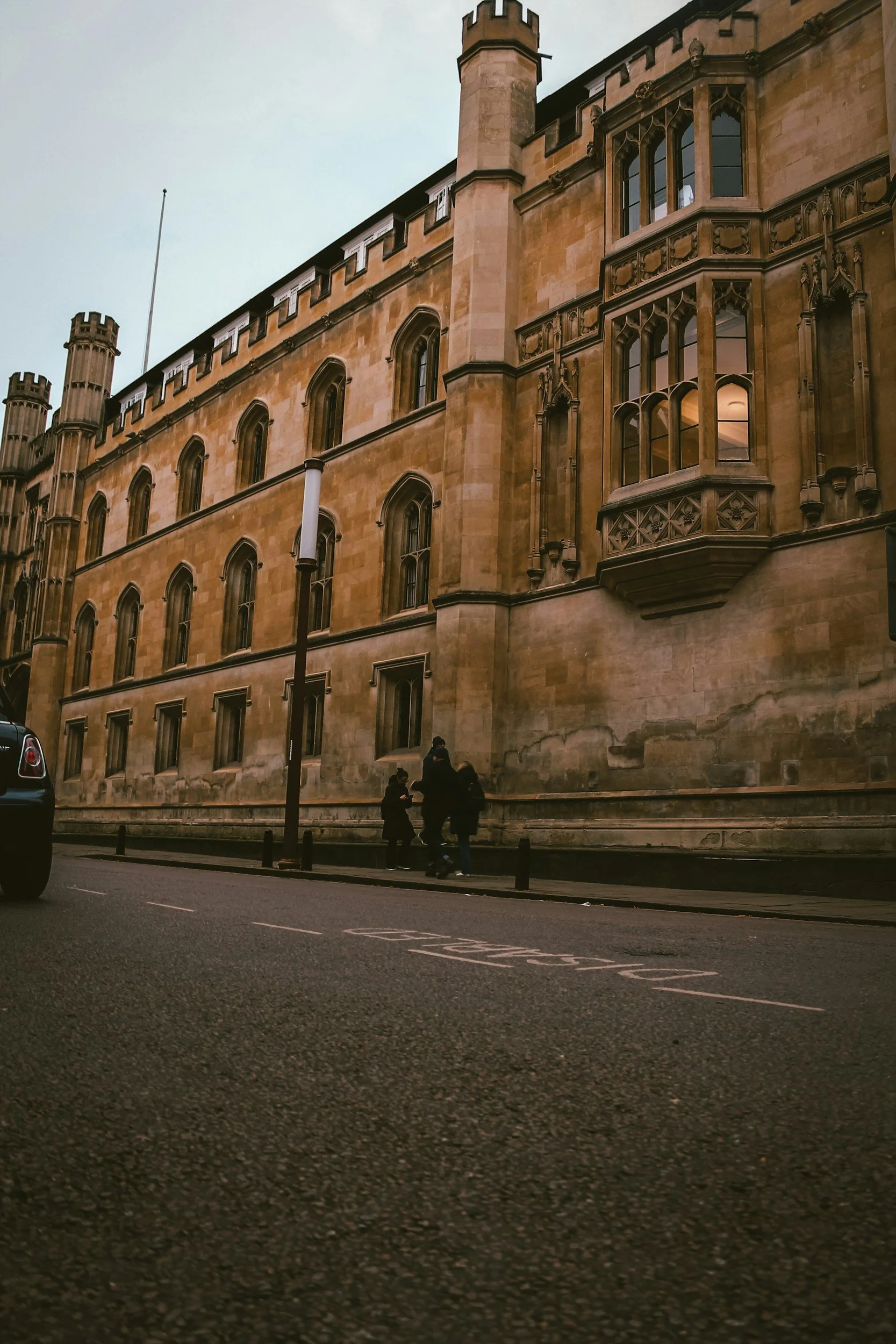 A historic stone building with ornate windows and towers, seen from a street with three people walking by and a dark car parked nearby.