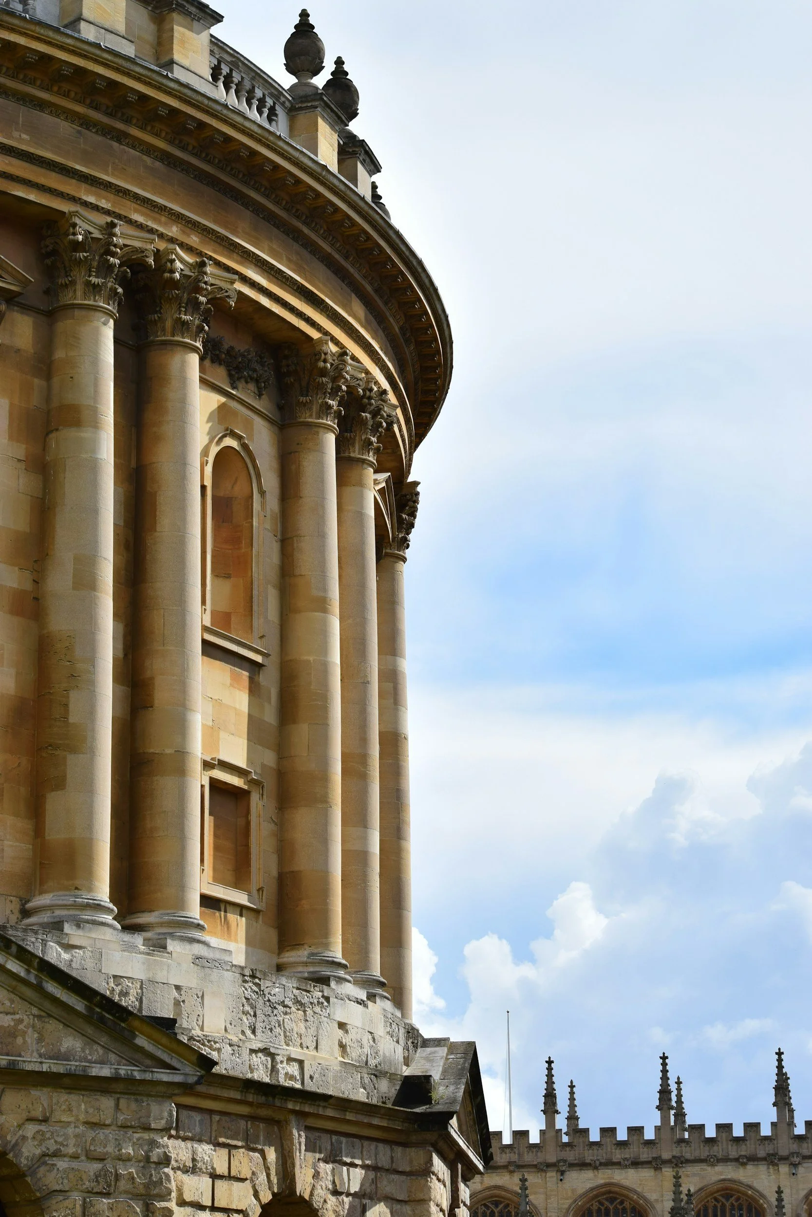 Close-up view of a historic sandstone building with classical columns and intricate architectural details. Part of a larger structure with battlements and spires visible in the background, set against a partly cloudy sky.