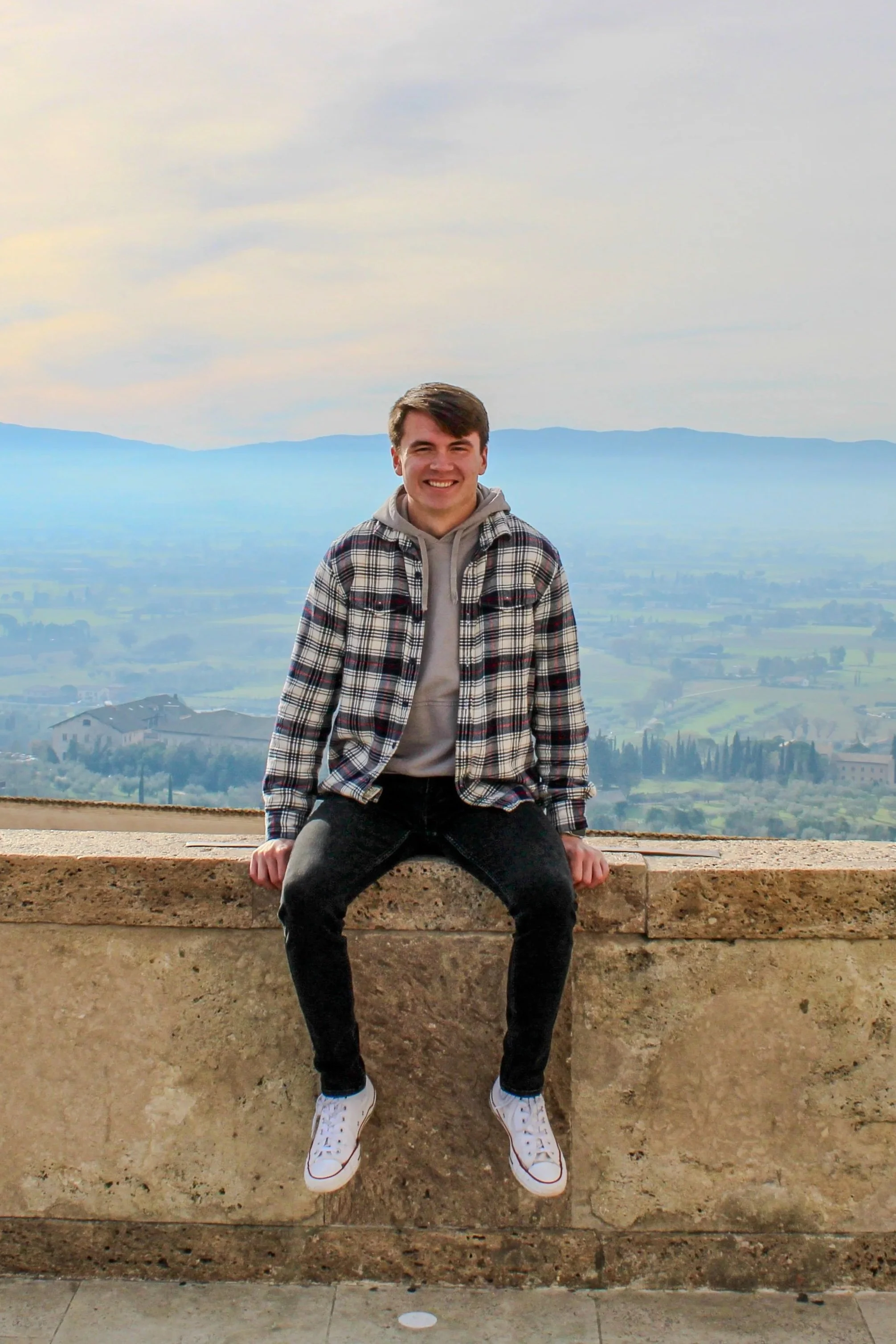 A young man sitting on a stone ledge with a scenic landscape of distant hills and a cloudy sky in the background.