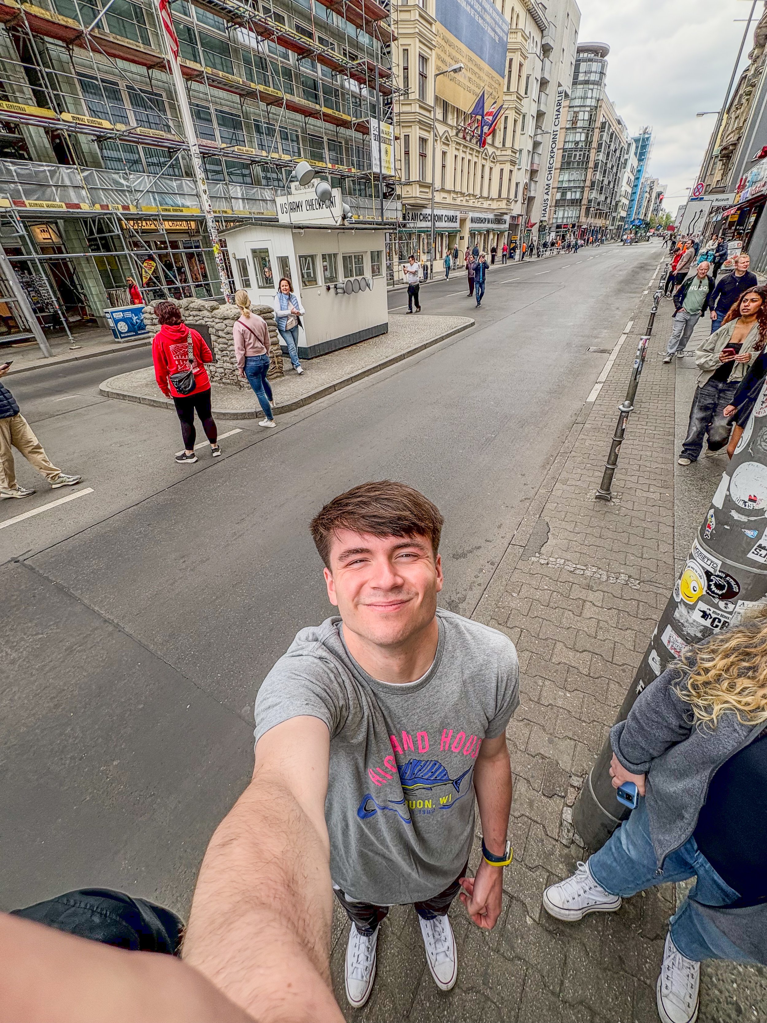Young man smiling taking a selfie on a city street with buildings, pedestrians, and scaffolding in the background.