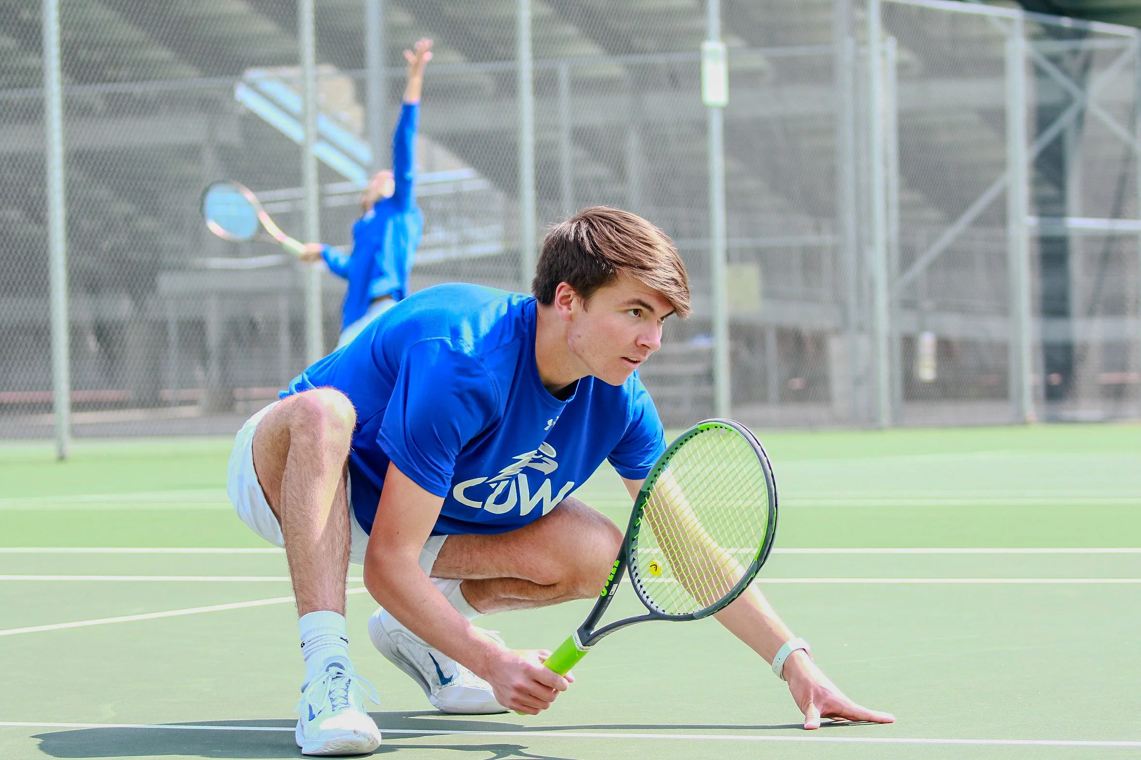 A young man in a blue t-shirt and white shorts crouches on a tennis court with a tennis racket, ready to hit a ball, while a woman in blue sportswear practices in the background.