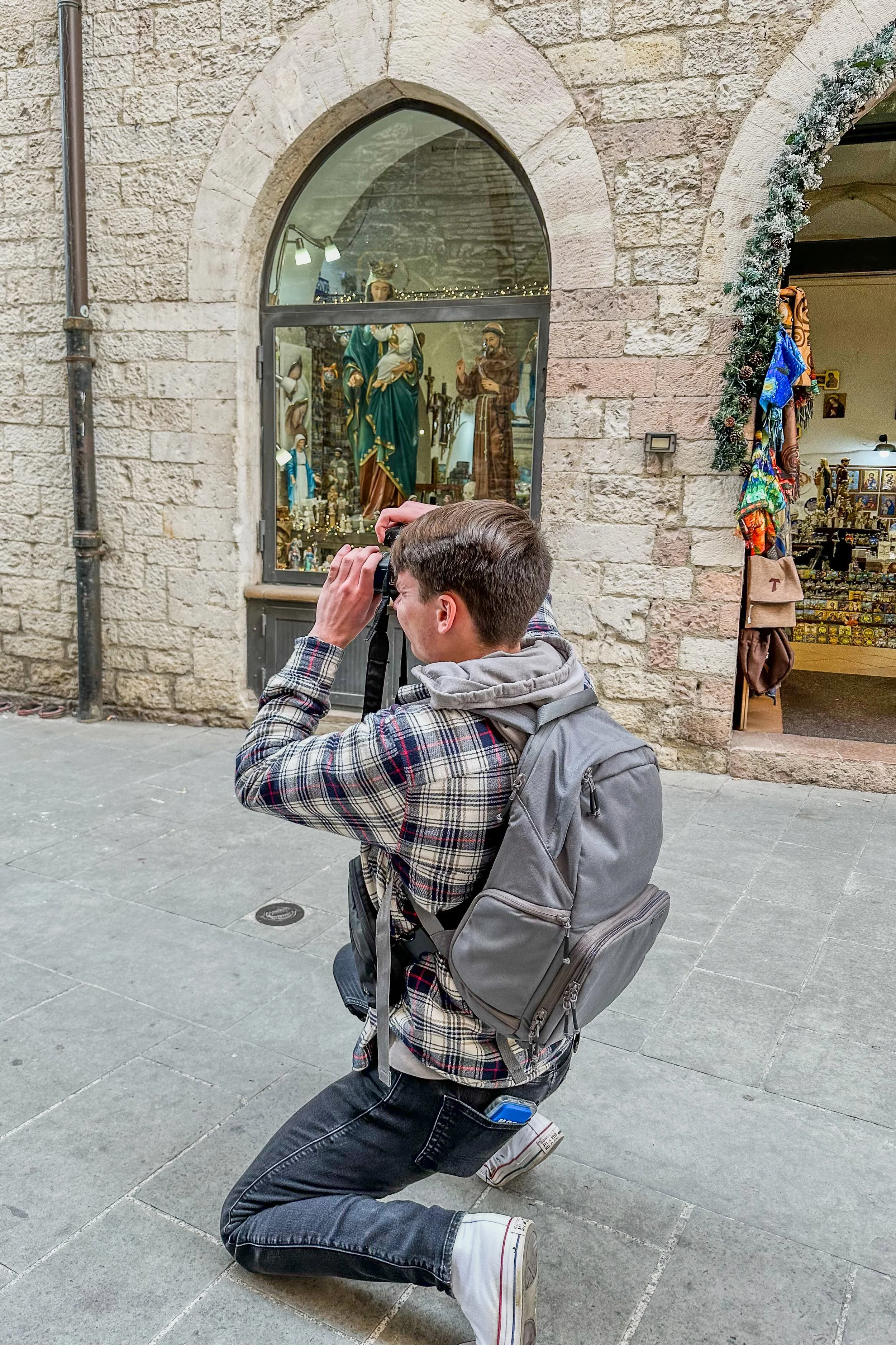 A young man kneeling on one knee with a camera taken a photo through a window display of religious statues.
