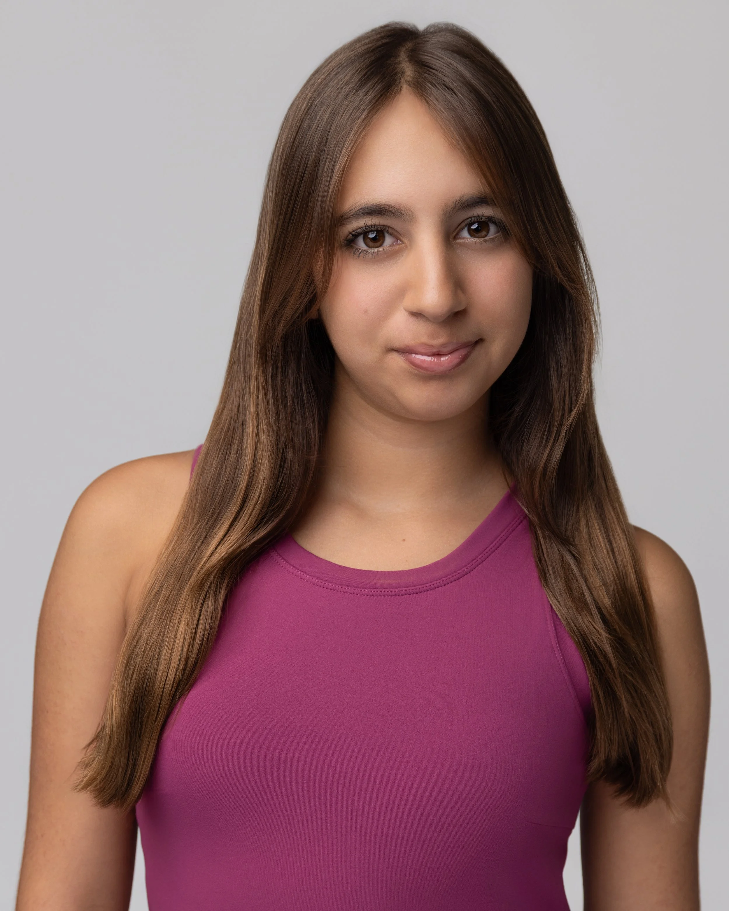 Layla Kazerouni, a young woman with long brown hair wearing a purple sleeveless top, smiling softly at the camera against a plain gray background.