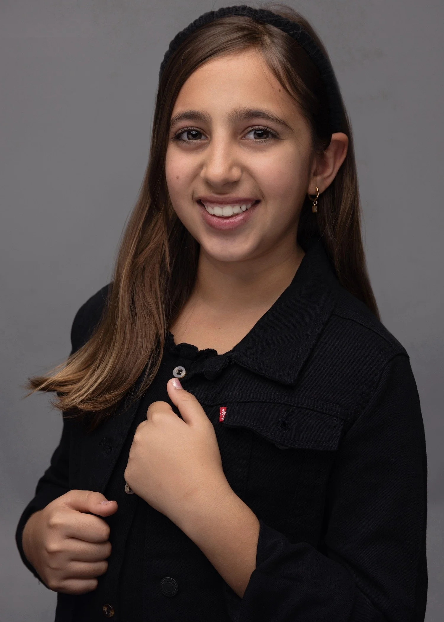 Layla Kazerouni, a young girl with brown hair, smiling, wearing a black jacket and a black headband, standing against a gray background.